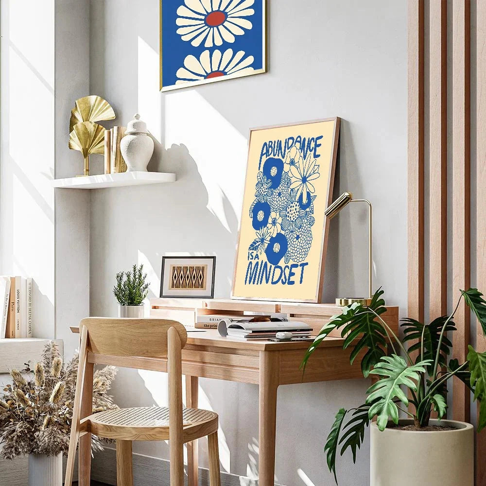 A blue and yellow floral art motivational poster titled "Abundance Mindset" displayed on a white rectangular table with a houseplant, surrounded by interior design elements like shelving, creating a serene and inspiring environment.