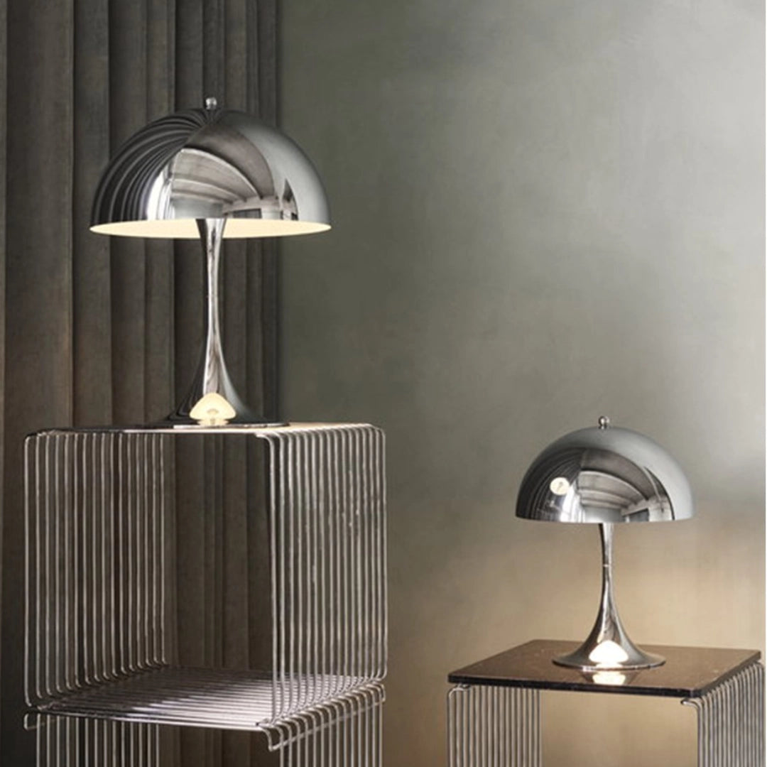 Adjustable gold dome desk lamp with reflective brass finish, placed on a wooden table in a stylish interior setting with grey walls and shelving.