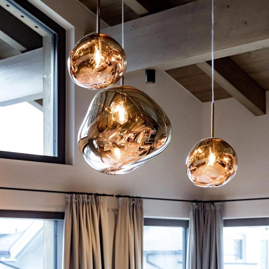Amber glass triple pendant light fixture with modern copper accents, hanging from the ceiling in a stylish interior setting. The warm light illuminates the space, complementing the room's wooden elements, a large window with curtains, and a tastefully designed wall.