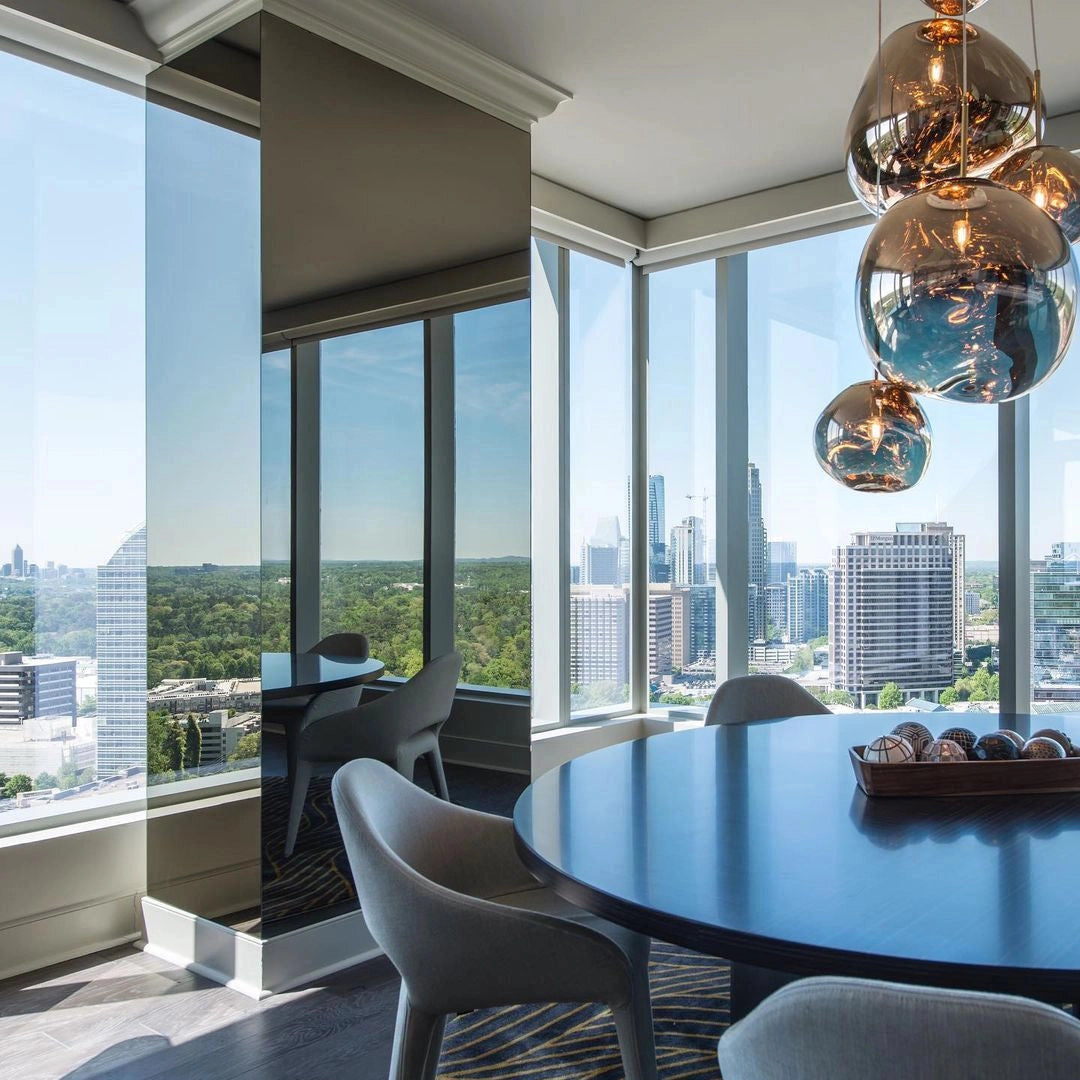 Modern copper ceiling lamp with an amber glass triple pendant design, suspended above a dining table in a bright room with large windows and views of the sky.