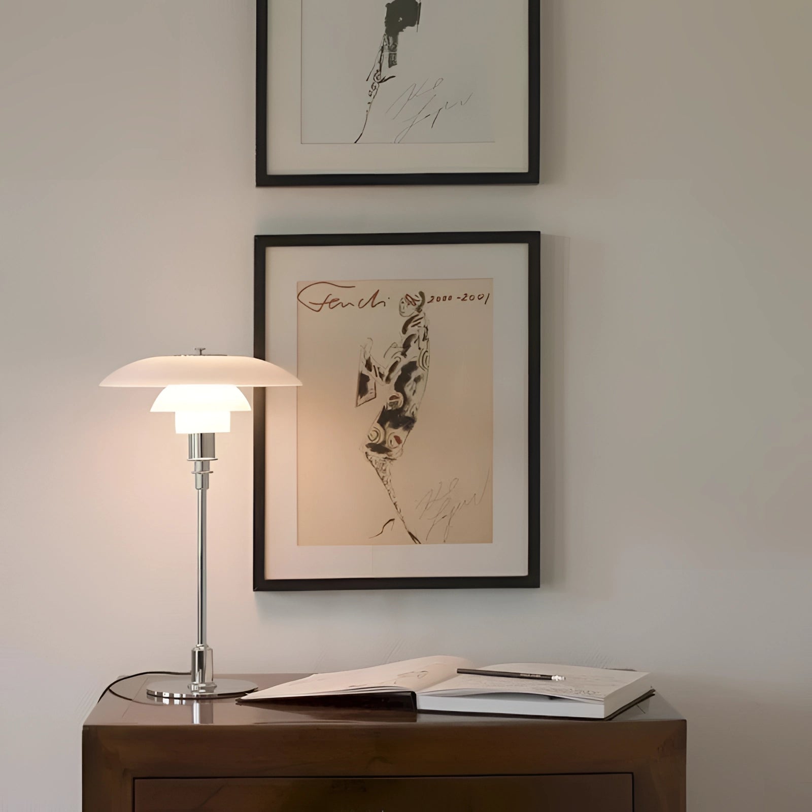 Minimalist brass table lamp with opal glass shade placed on a wooden table beside a wall adorned with picture frames.