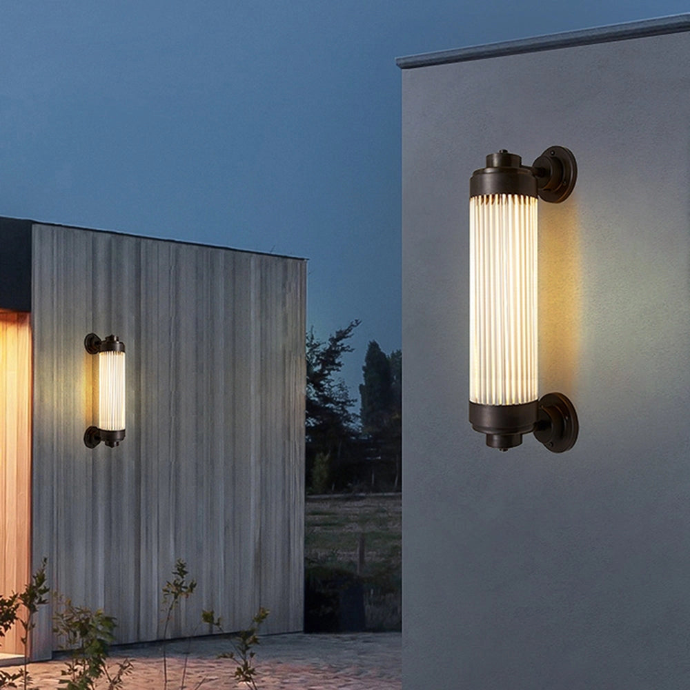 Bronze vintage glass tube wall sconce with industrial design, featuring a large 25.2-inch size, mounted on a wall next to a plant and a wooden accent, providing ambient lighting against a soft sky backdrop.