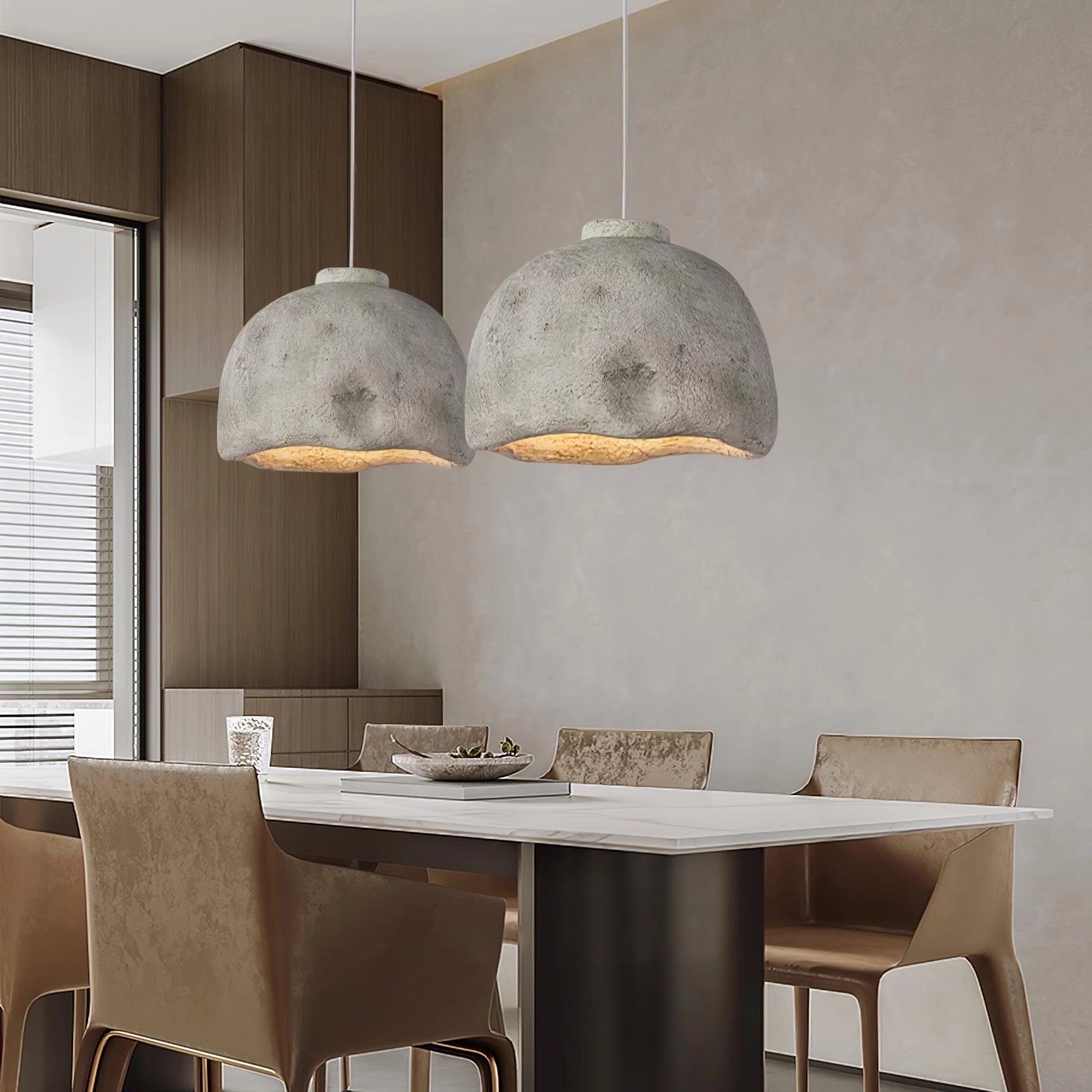 Concrete pendant light hanging above a wooden kitchen and dining room table, surrounded by chairs, showcasing an industrial and minimalist interior design with a grey color scheme.