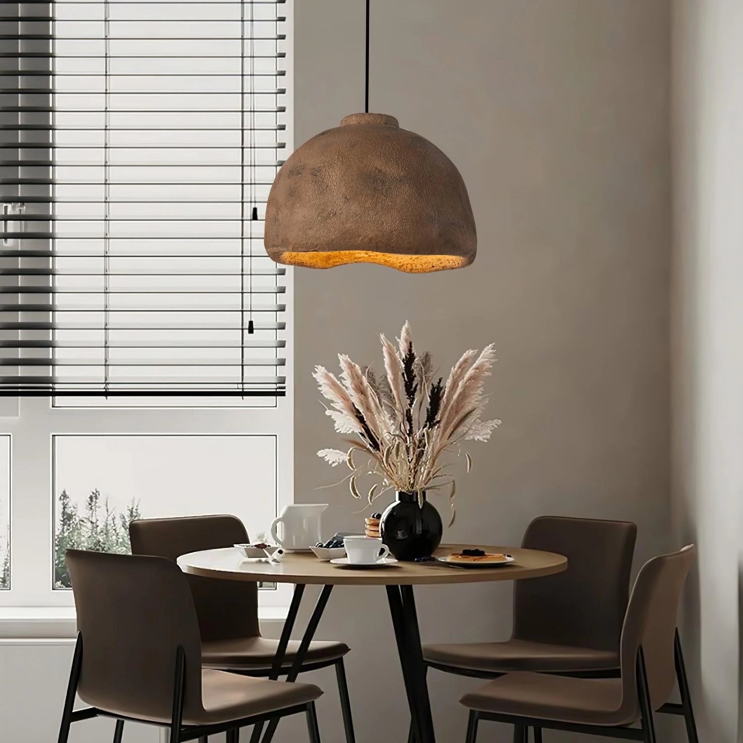 Concrete pendant light hanging above a wooden table surrounded by chairs, with indoor plants and textile elements adding a touch of nature to the minimalist and eco-friendly interior design setting.