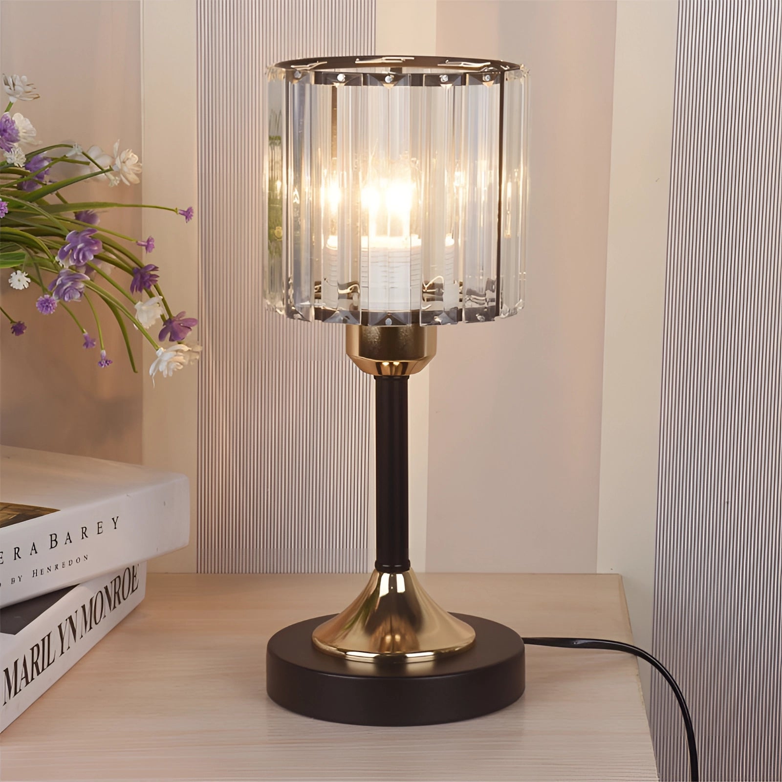 A modern bedside lamp with a crystal and brass base, featuring a gold finish, and a black round lampshade. The lamp is positioned on a wooden table, surrounded by contemporary interior design elements such as a potted plant and hardwood flooring.