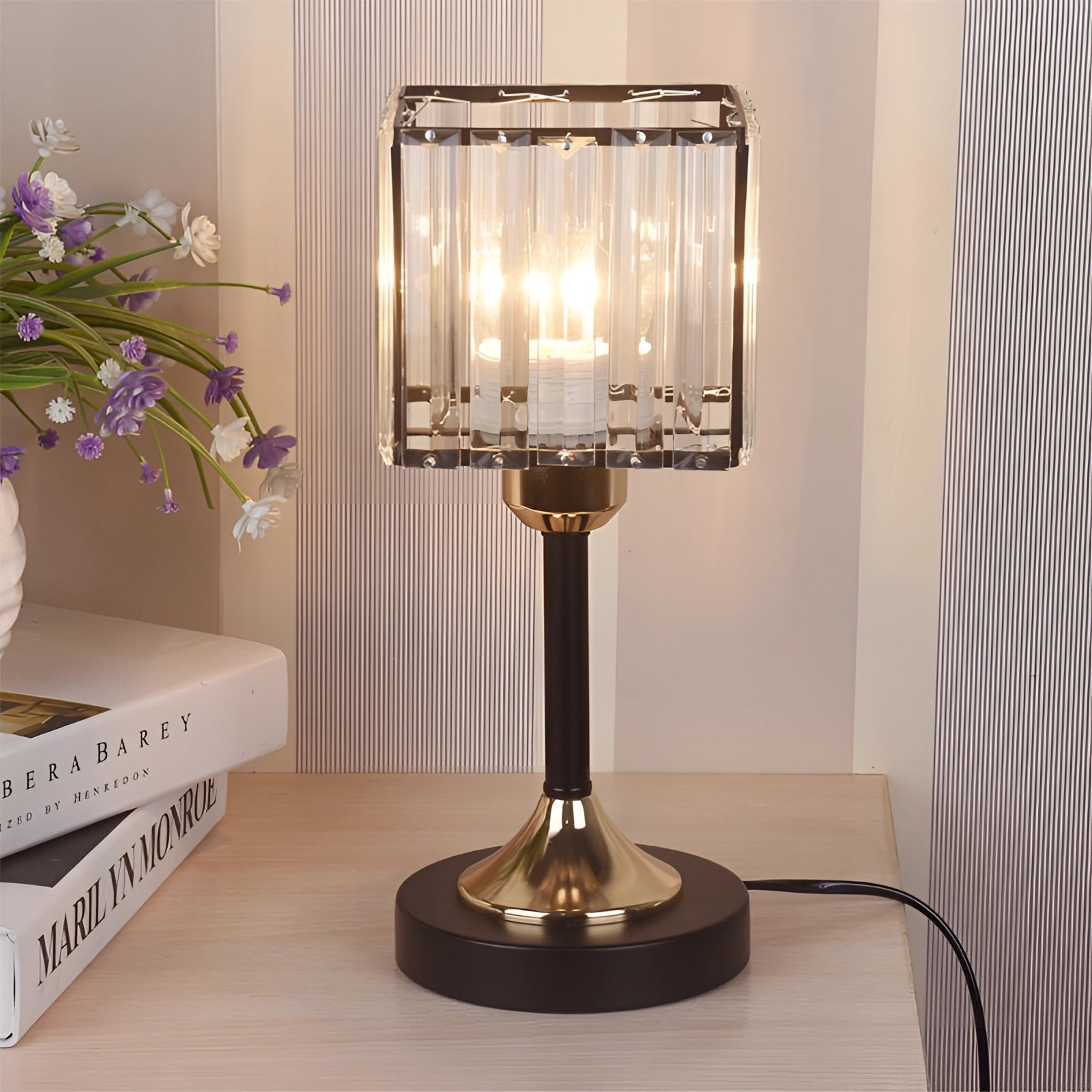 A modern bedside lamp with a square black shade and a gold-finished crystal brass base, showcased in an interior setting with wooden flooring.