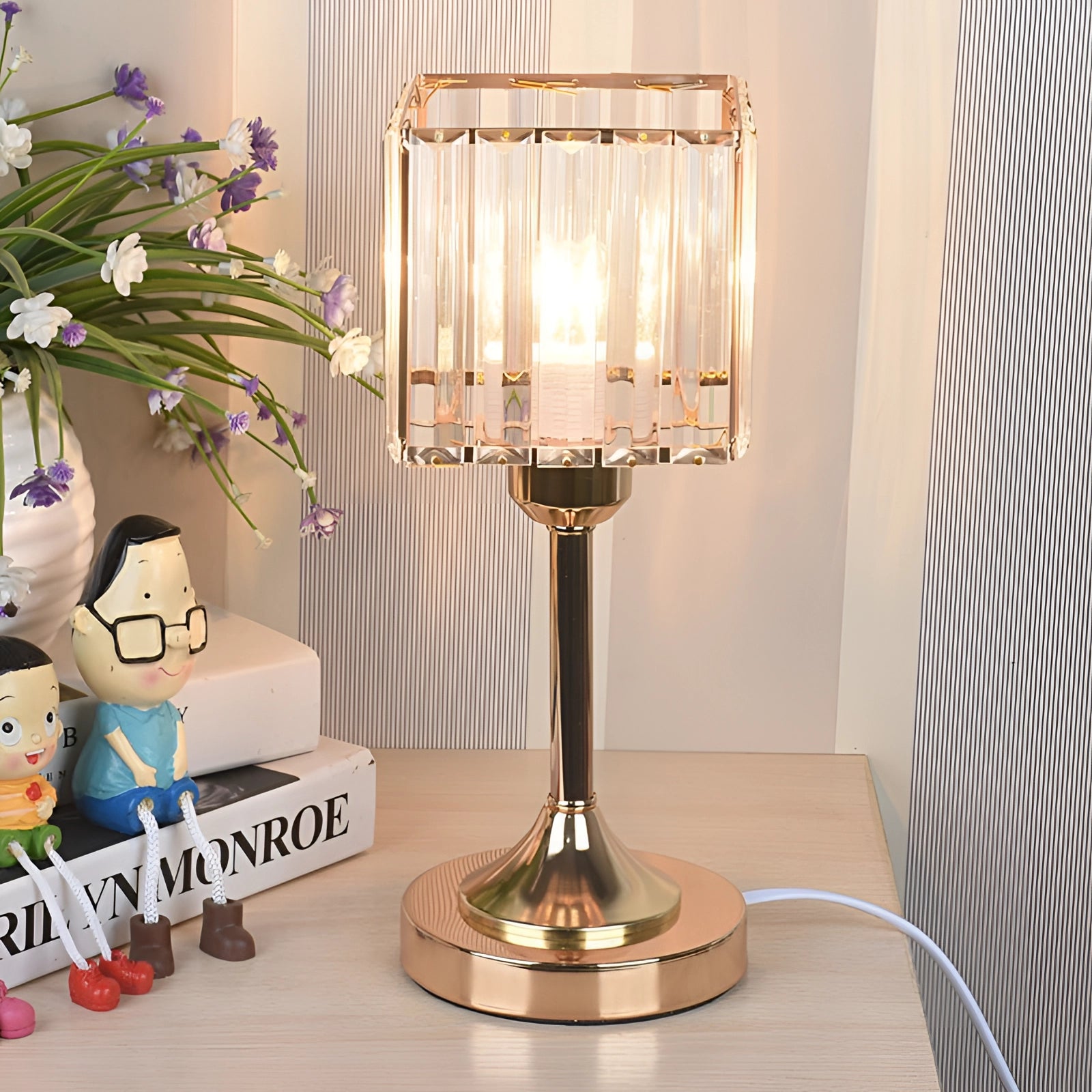 Gold finish crystal brass table lamp with a square base, placed on a wooden surface, elegantly illuminating a modern interior space with decorative plants nearby.