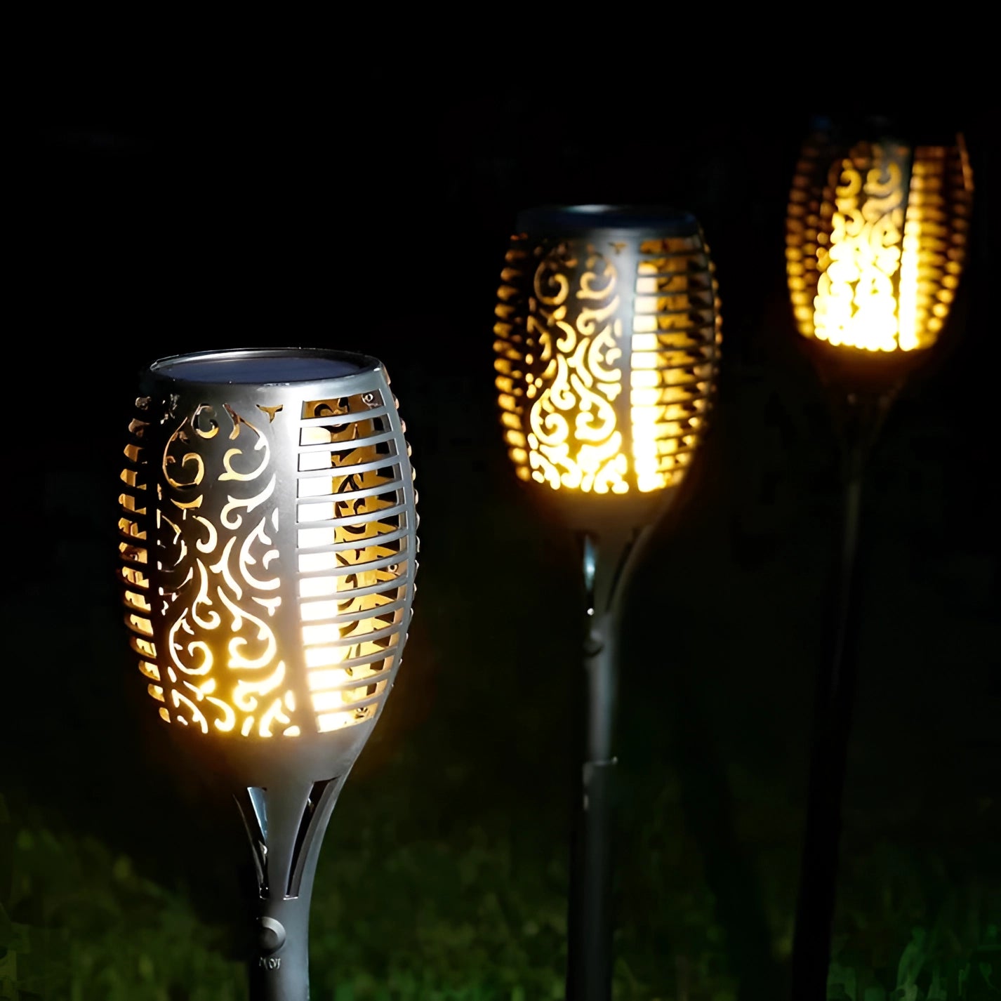 Decorative LED solar flame torches with a tribal design, waterproof, illuminating a garden area with a warm amber glow.