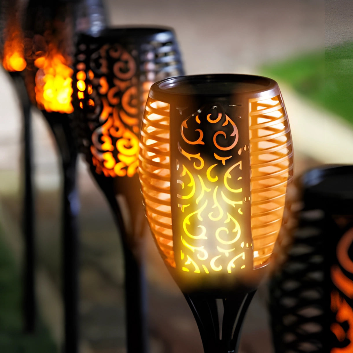 Decorative LED solar flame torches glowing with amber and orange light, designed with a tribal pattern, standing outdoors in a garden setting, showcasing their large size.