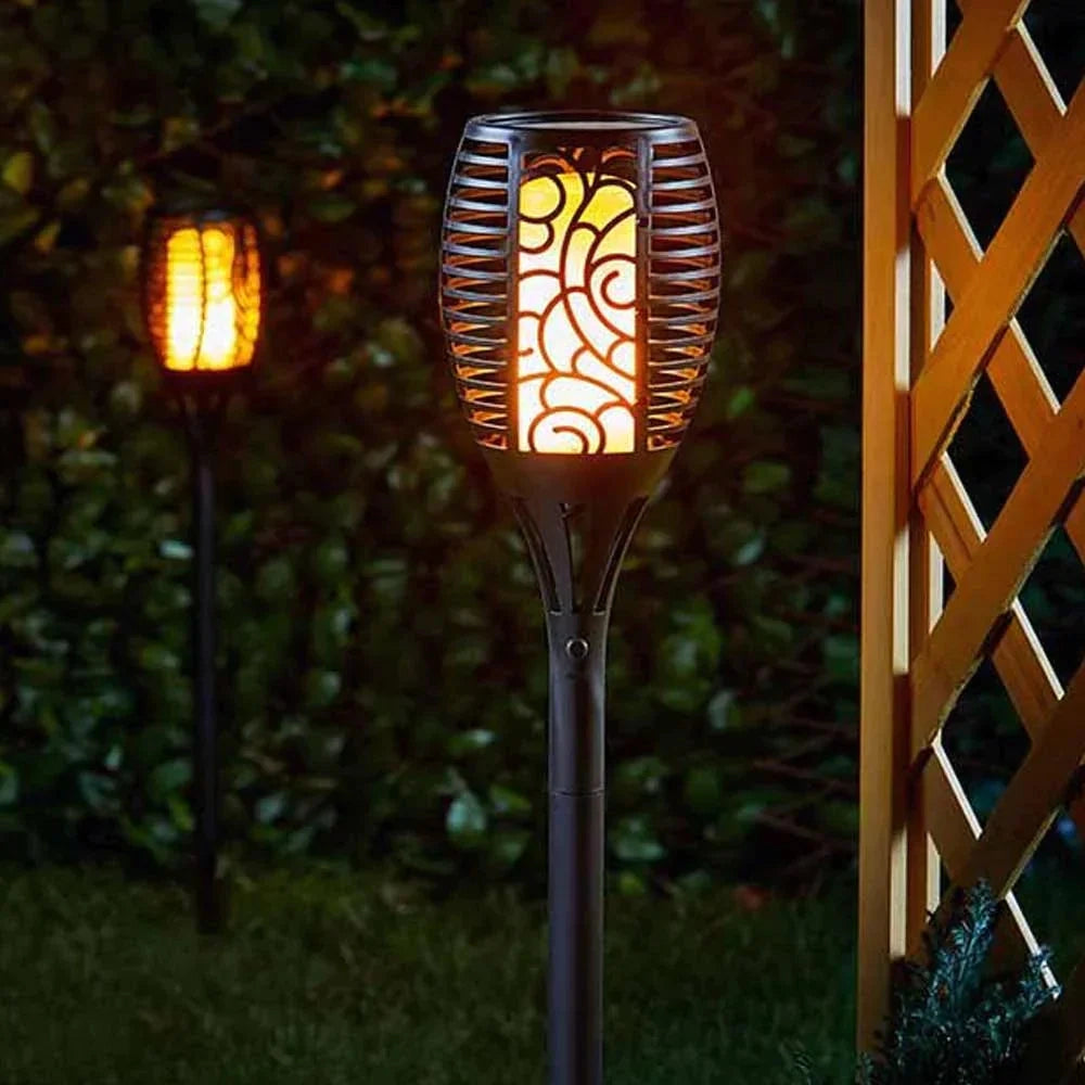 Decorative LED Solar Flame Torches illuminating a garden with a warm amber glow, surrounded by grass and plants, featuring waterproof tribal designs.