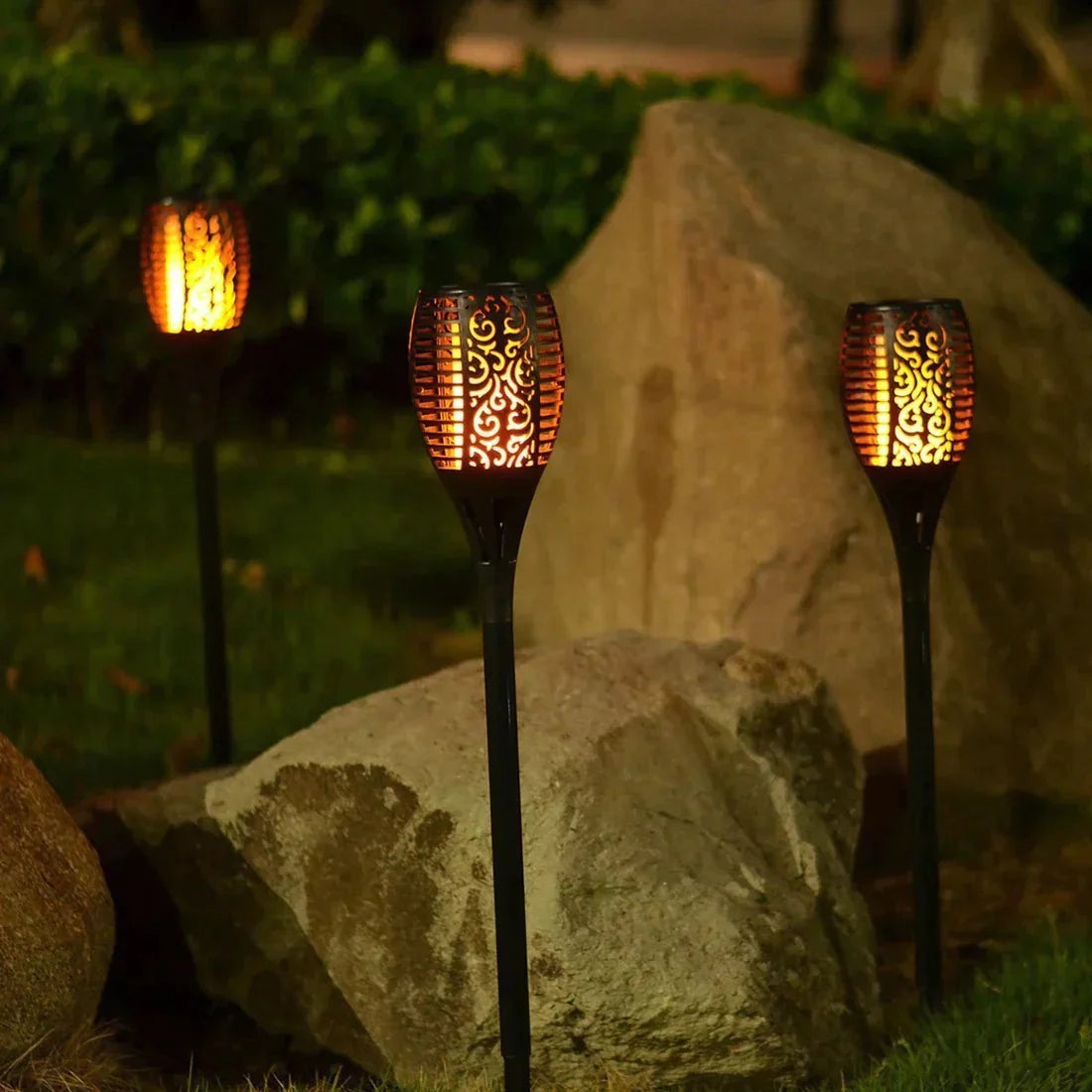 Decorative LED solar flame torches illuminating a garden at night with a warm amber glow, surrounded by grass.