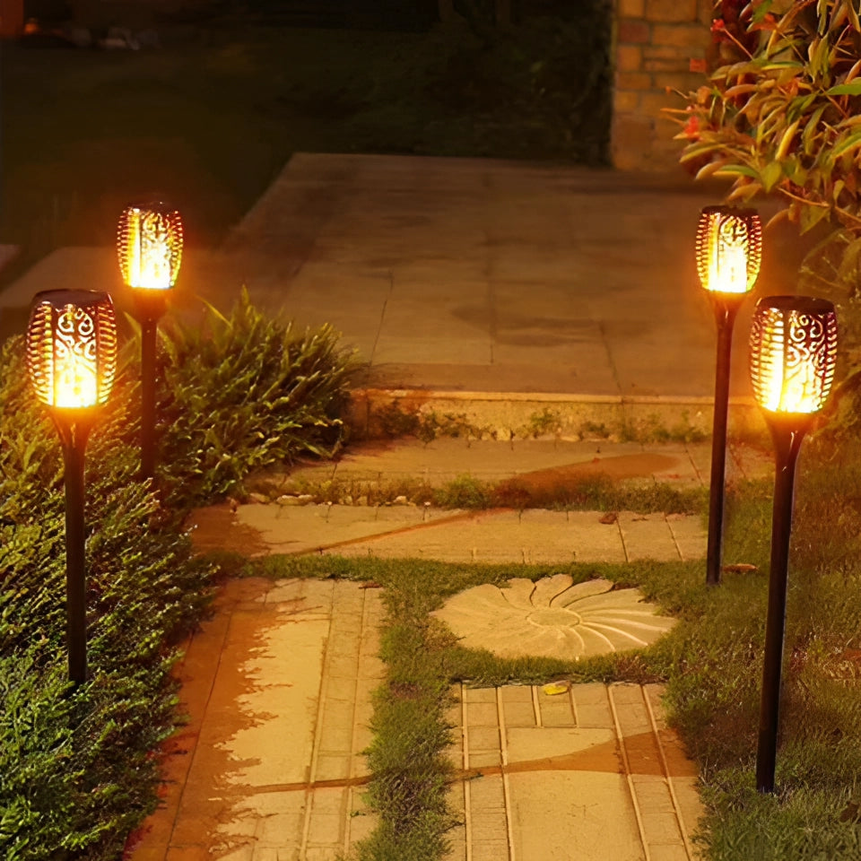 Decorative LED solar flame torches illuminating a garden area with vibrant amber light, surrounded by plants and casting shadows on the road surface.