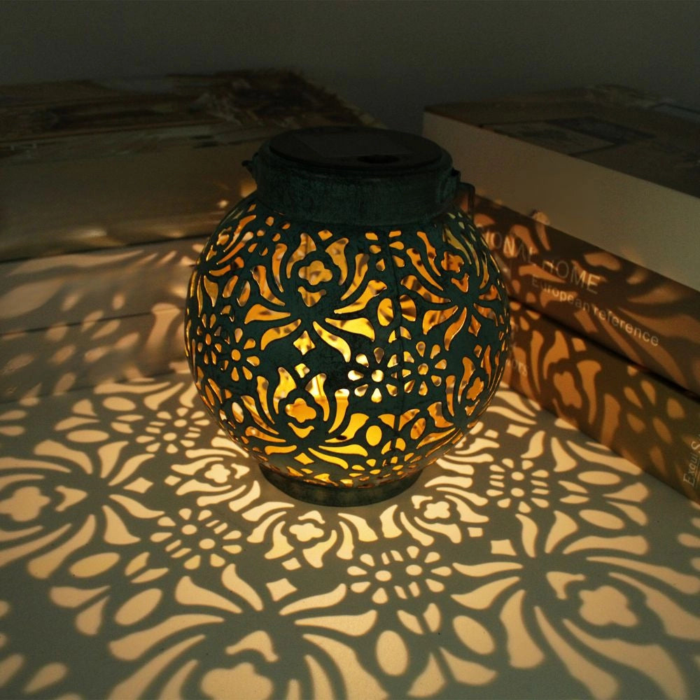 Decorative solar lantern with floral cut-out design, casting intricate shadows, featuring an amber tint and symmetrical circular patterns, suitable for ambient outdoor lighting.