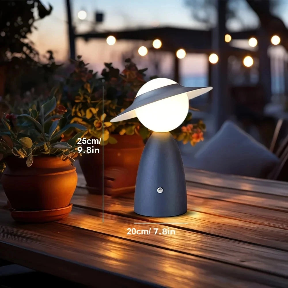 A modern UFO-shaped table lamp with a mushroom design, placed on a wooden table next to a houseplant and a flowerpot, with a window and tree visible in the background.