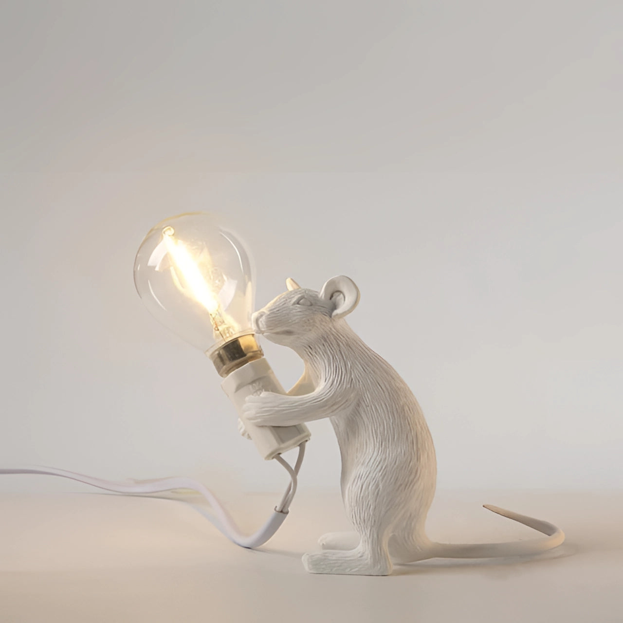 Sitting white gold mouse-shaped lamp featuring a light bulb held in its paws, with intricate metallic details and a visible power cable, designed as an eclectic piece of decor.