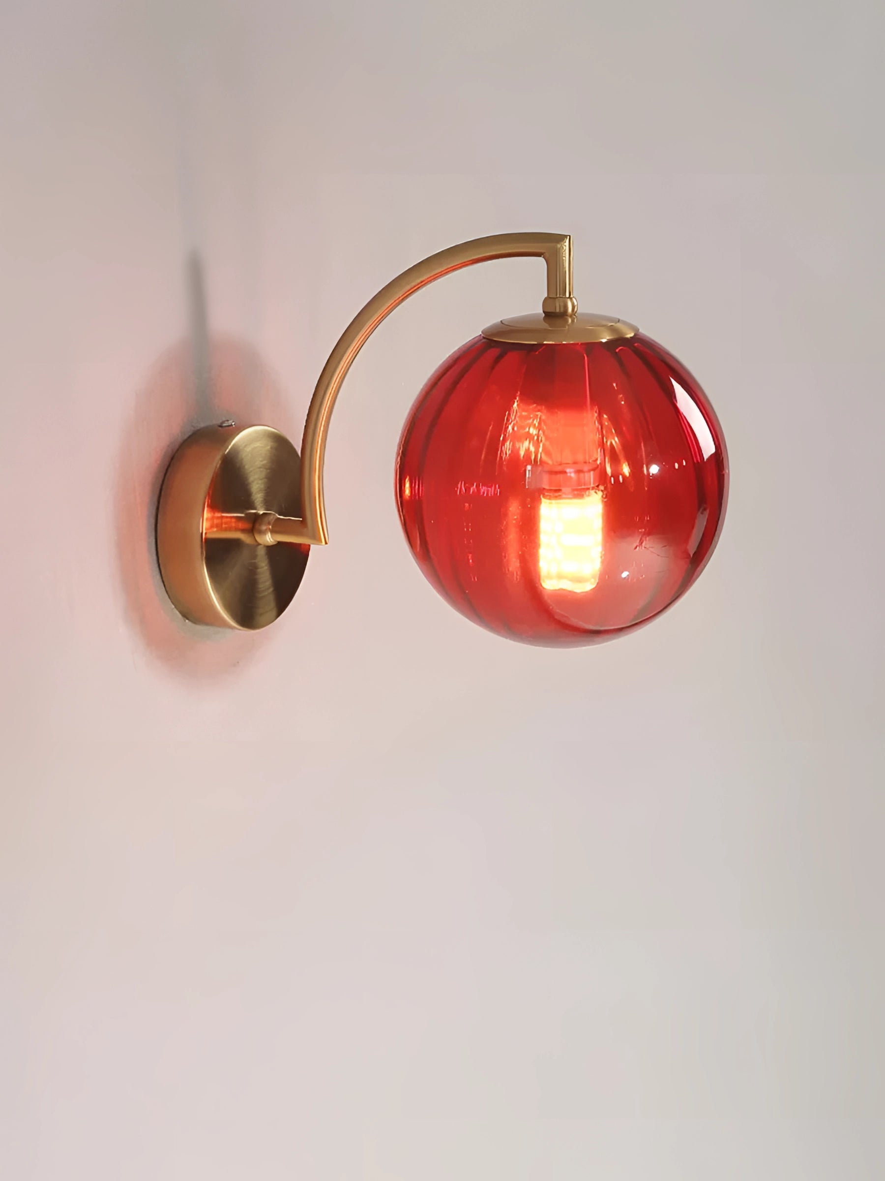 Modern brass wall sconce with ribbed glass globe, red variant, featuring a sleek metal design and mounted against a blank background.