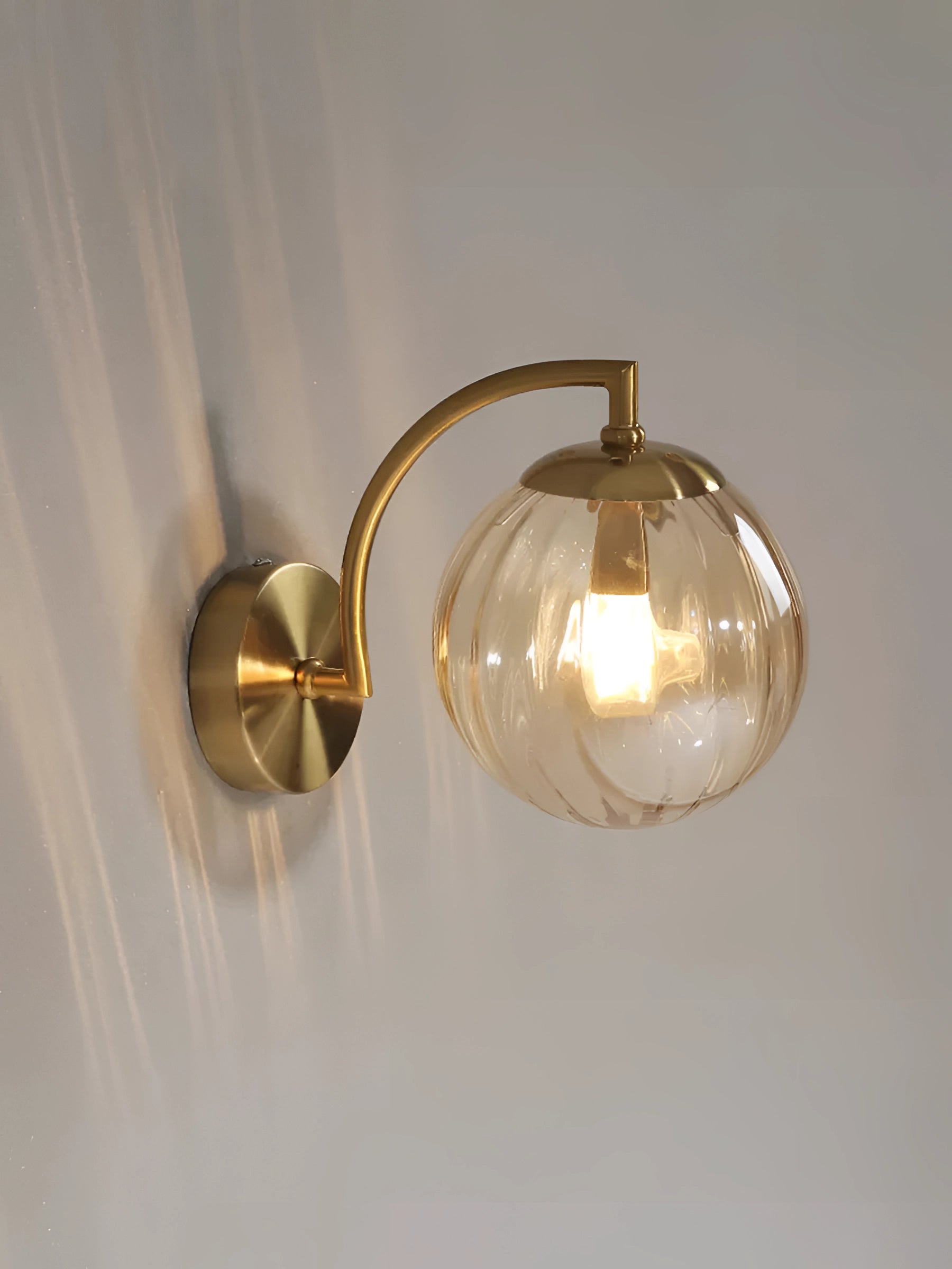 Modern brass wall lamp with ribbed glass globe in amber finish mounted on a wooden wall, illuminating a warm light.