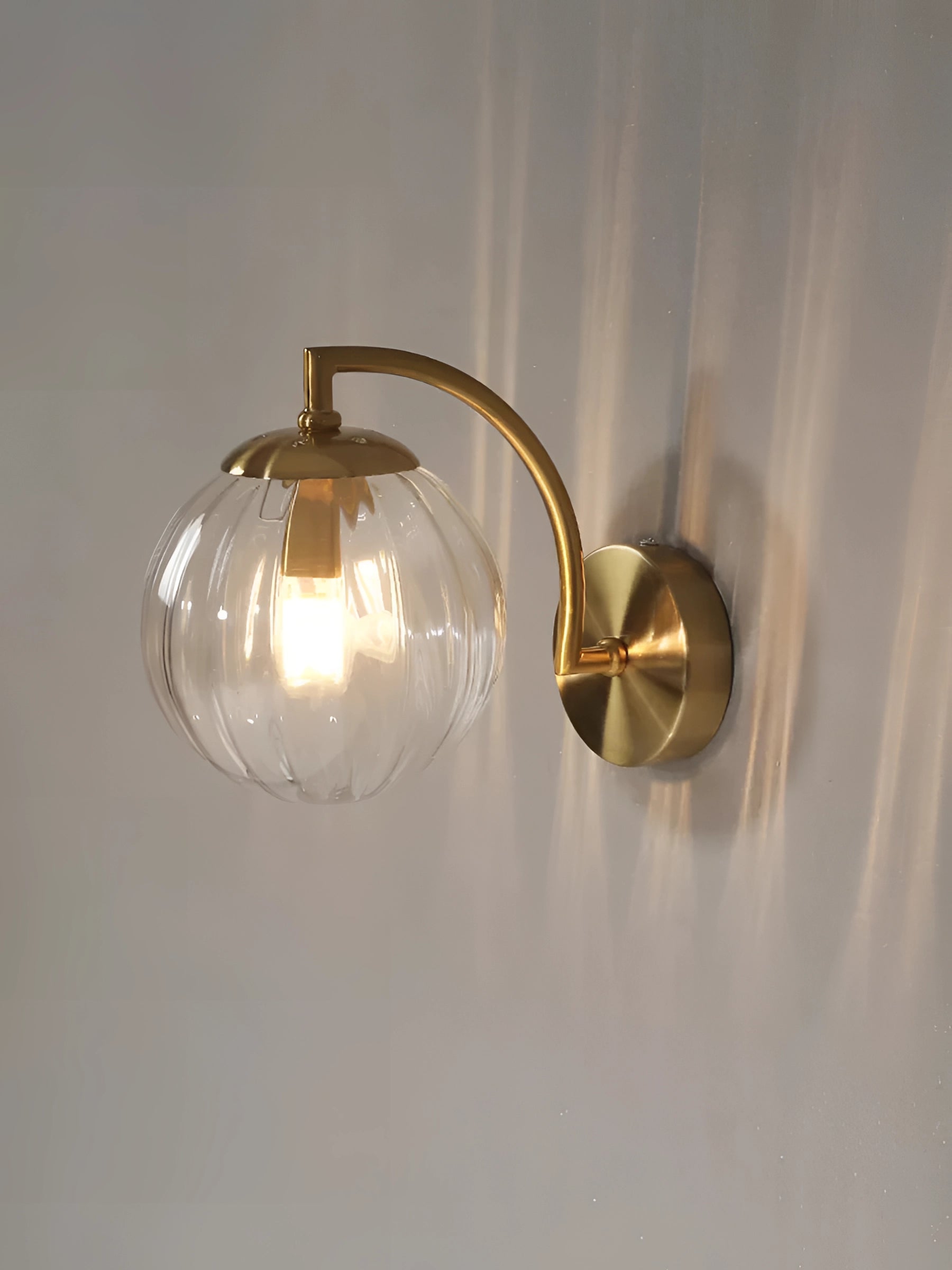 Modern gold wall sconce with ribbed transparent glass globe and brass fixture, mounted on a wooden wall.