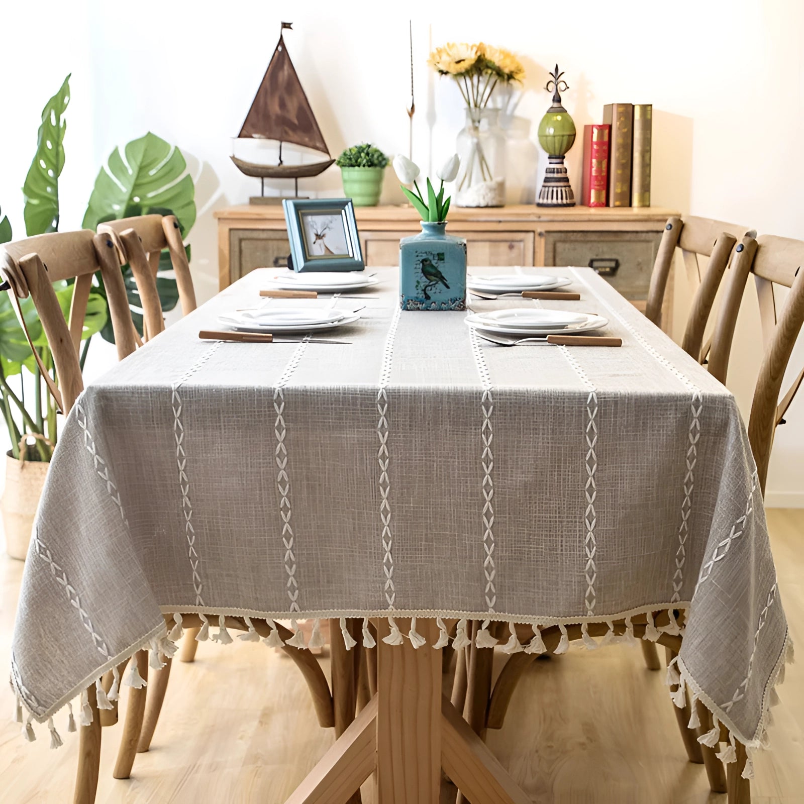 Light brown rectangular table with a grey linen tassel tablecloth in an eco-friendly Scandinavian boho design, accompanied by a chair and a small plant, set in a well-lit interior space.