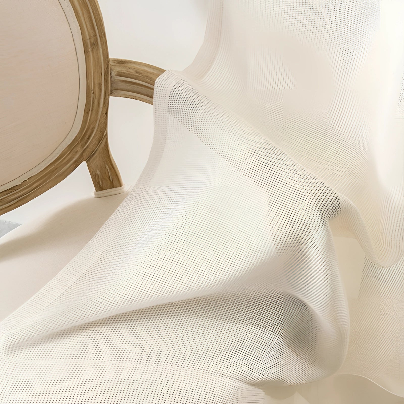 Ivory sheer curtains hanging elegantly in a living room, allowing soft natural light to filter through, complemented by a wooden table and chair in the foreground.