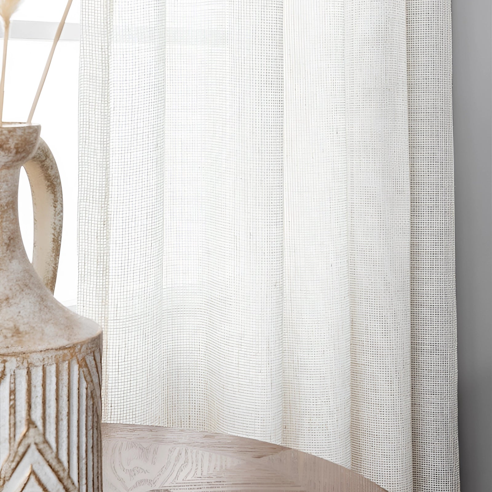 Ivory sheer curtains elegantly hanging in a living room with beige and grey furniture, including a chair and a couch, alongside a wooden table.
