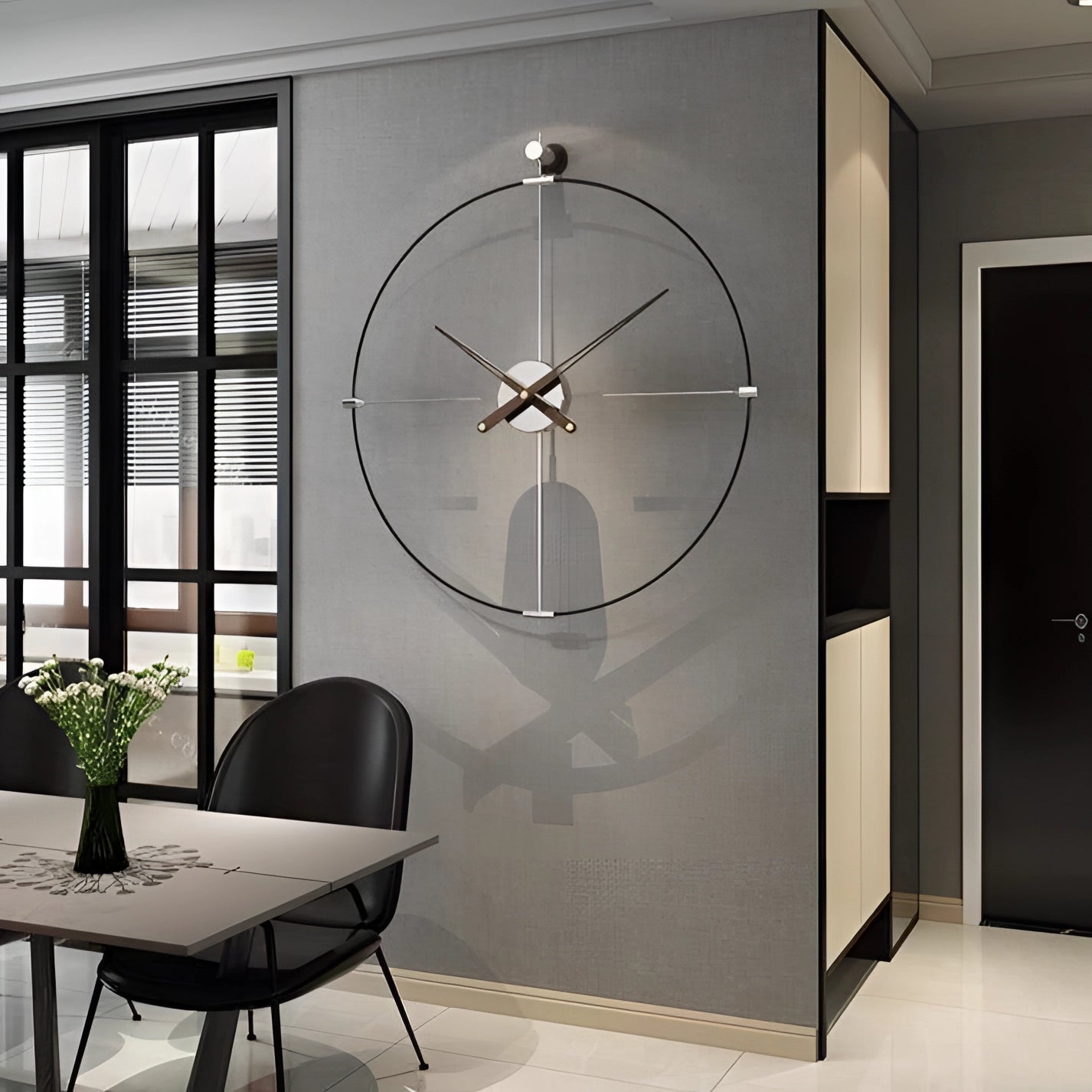 Large black luxury metal wall clock with a modern minimalist design, measuring 63 x 69 cm, mounted on a wall in an interior setting, surrounded by furniture, a wooden table, and houseplants in flowerpots near a window.