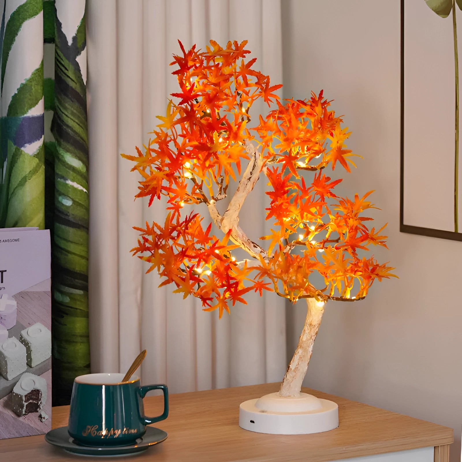 LED Maple Bonsai Tree Lamp with orange maple leaves, illuminated and positioned near a window. The lamp is USB powered, designed to resemble autumn foliage, with branches and twigs creating a cozy atmosphere. Nearby, a coffee cup rests on a textile surface, enhancing the warm decor setting.