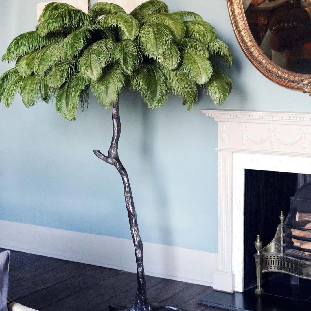 A luxury feather floor lamp designed to resemble a gold palm tree, standing next to a green houseplant on a shelf, showcasing an art deco style.