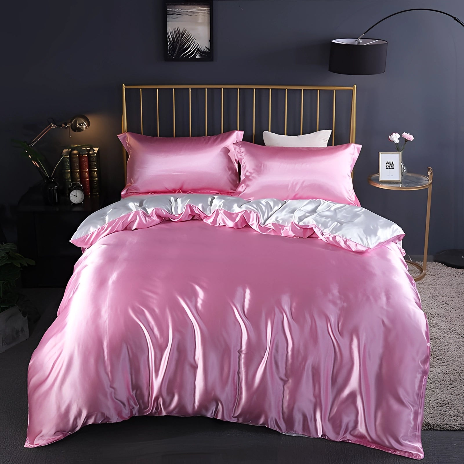 A luxurious silk bedding set in pale pink color, displayed on a neatly made bed in a well-lit bedroom. The set includes fitted sheets, and the room features a window allowing natural light, a wooden lamp, and a potted plant, all contributing to a cozy interior design.