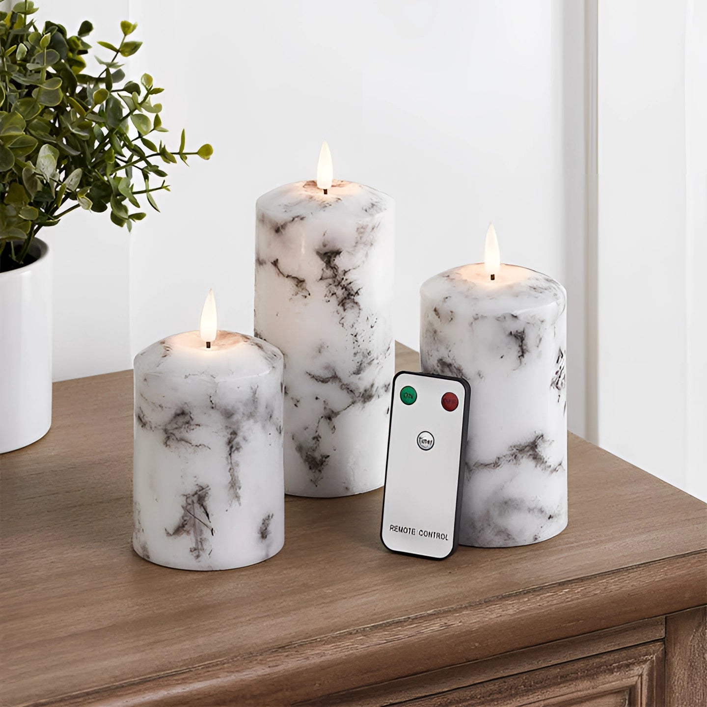 Marble design flameless LED candles on a wooden cabinet, surrounded by potted plants and decorative items, with a remote control visible nearby.