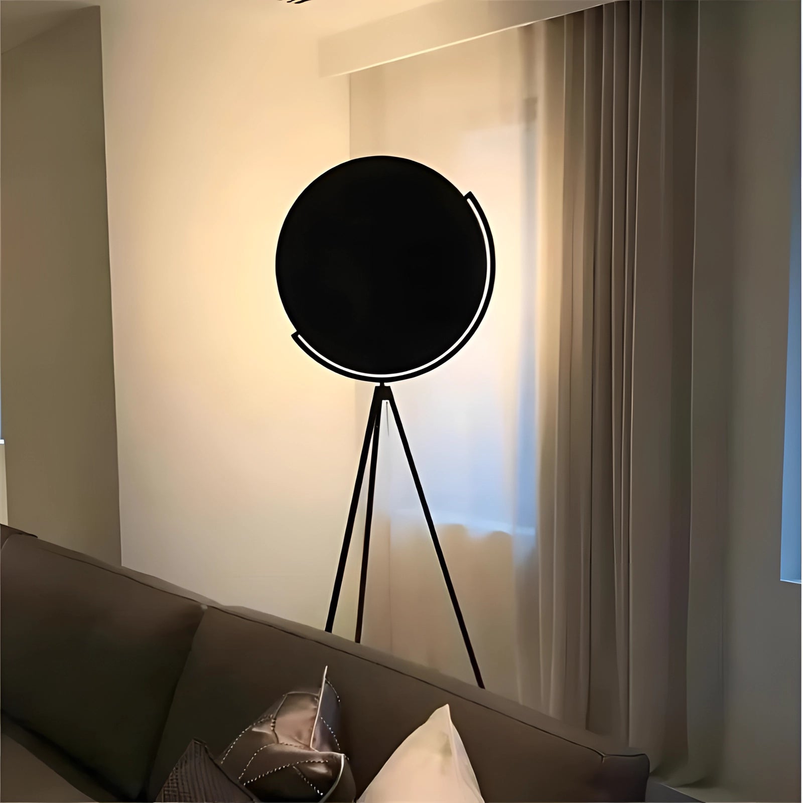 A modern black tripod floor lamp with an adjustable minimalist design, featuring a sleek black shade and wooden tripod legs, standing in a stylish interior setting with a couch, window, and contemporary artwork.