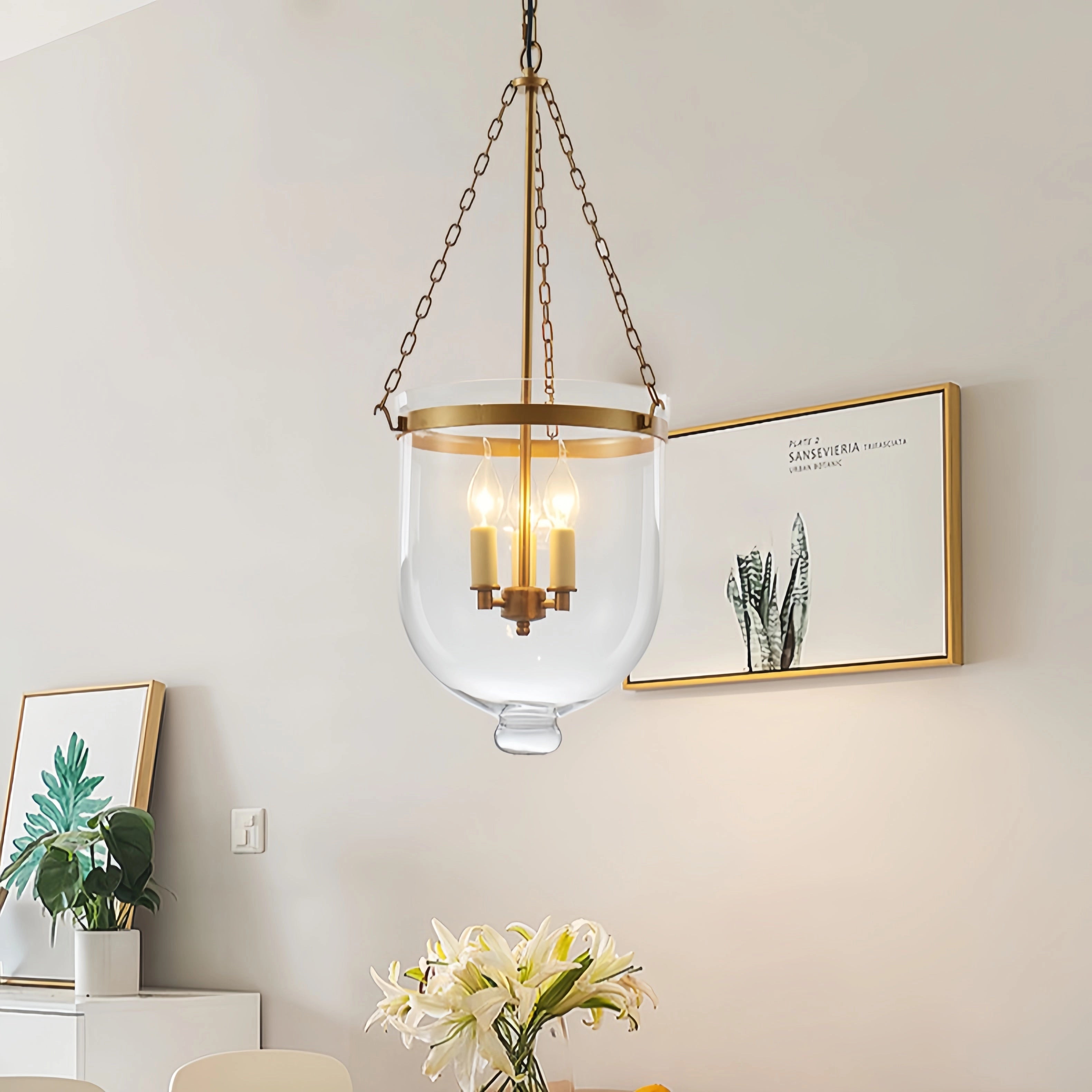 Modern brass glass pendant light with a luxurious gold finish, displayed hanging in an elegantly designed indoor setting, featuring wooden and yellow accents, plants, and picture frames in the background.
