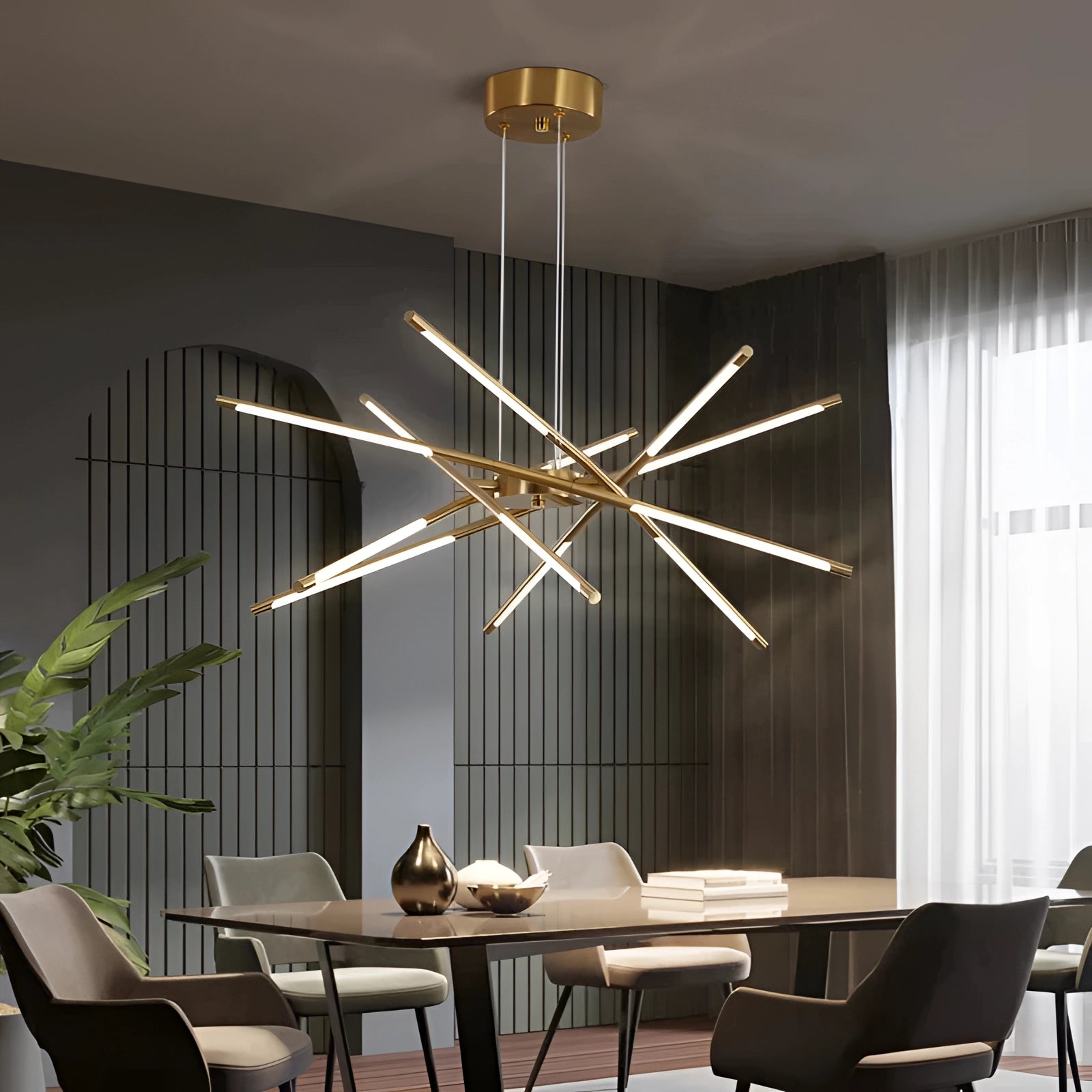 Modern brass LED chandelier illuminating a luxury dining room setting with a wooden table, chairs, plant decor, and curtains in the background.