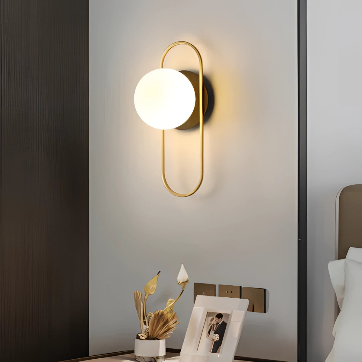 Modern dual globe brass wall sconces in warm white and gold finish, mounted on a white wall in a minimalistic interior setting, with nearby plants and wooden elements adding to the decor.