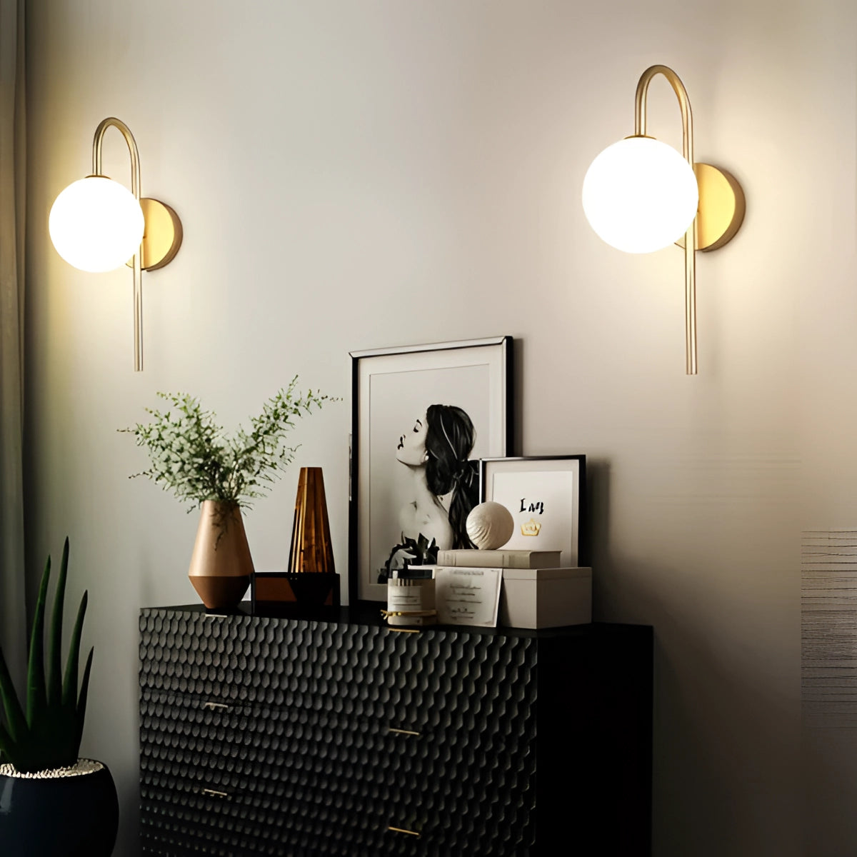 Modern dual globe brass wall sconces mounted on a grey wall with minimalistic design, accompanied by a houseplant in a flowerpot, wooden elements, and a picture frame in a contemporary interior setting.