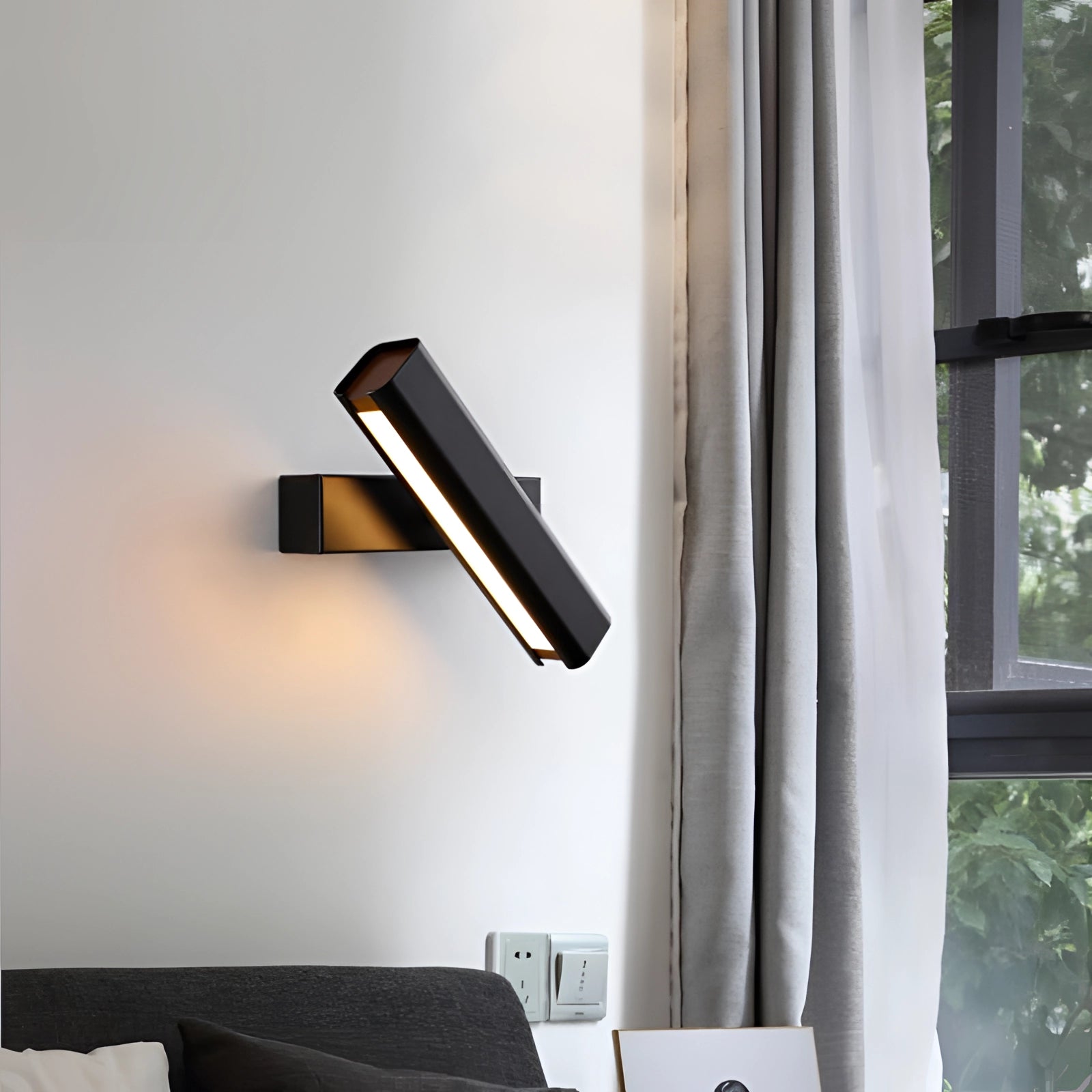 Modern LED wall sconce in black and gold finish, mounted on a sleek interior wall with contemporary design elements including a couch, window, and wooden accents, providing warm white illumination.