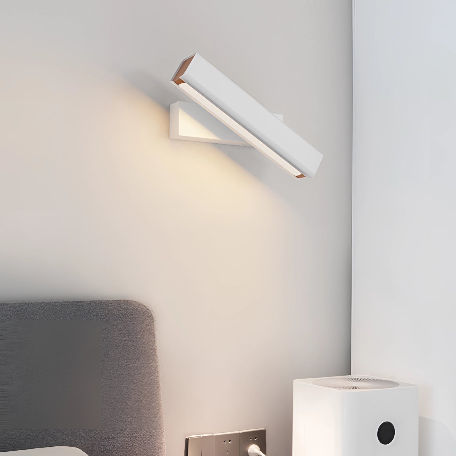 Modern LED wall sconce in black and gold with a white finish, emitting warm white light, mounted on a wall in a contemporary interior setting with a grey couch and wooden elements.
