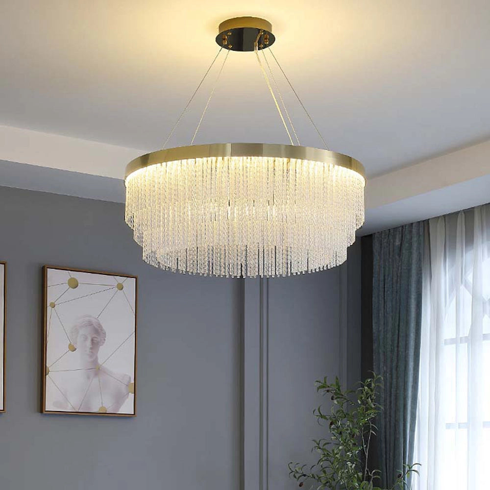 Modern luxury circular crystal LED chandelier with a brass finish, measuring 23.6 inches, elegantly hanging in a sophisticated interior setting. The room features a large window with curtains, wooden elements, and plants, creating a stylish and well-lit ambiance.