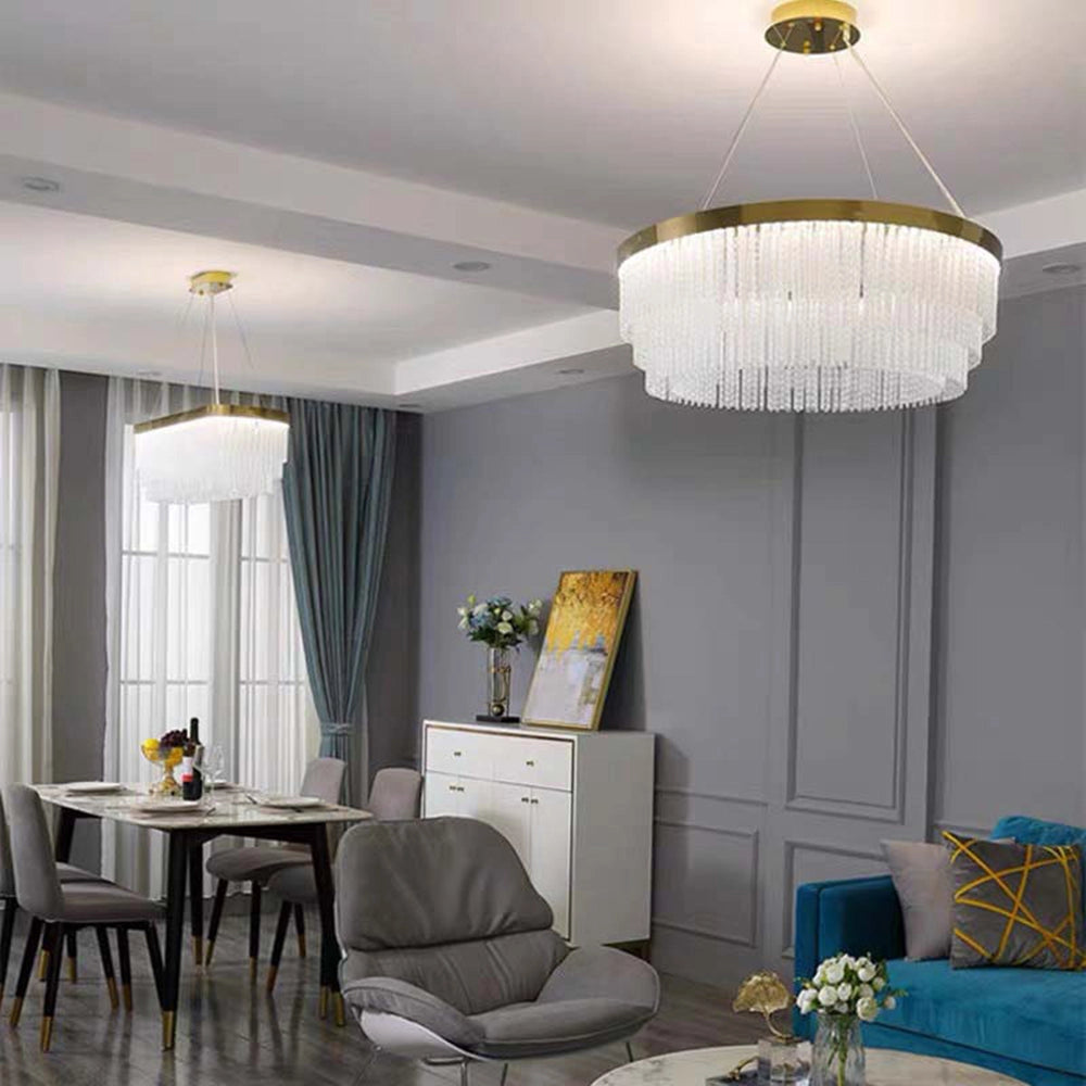 Modern luxury circular crystal LED chandelier with a brass finish hanging in a stylish interior setting, surrounded by contemporary furniture including a table, chairs, and a couch near a window.