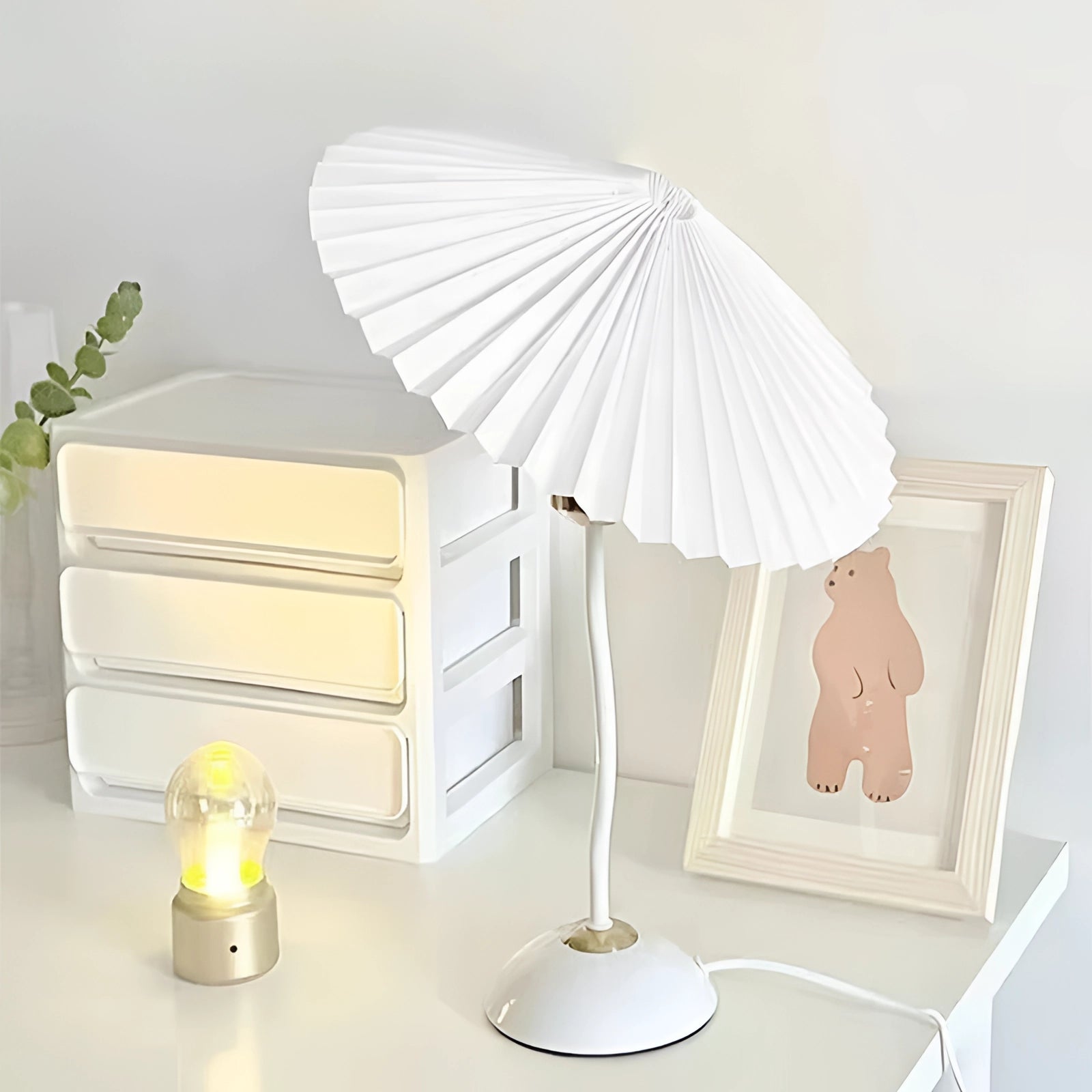 A modern pink origami table lamp with a pleated accordion design, displayed with a white variant, positioned on a wooden floor next to a green plant.