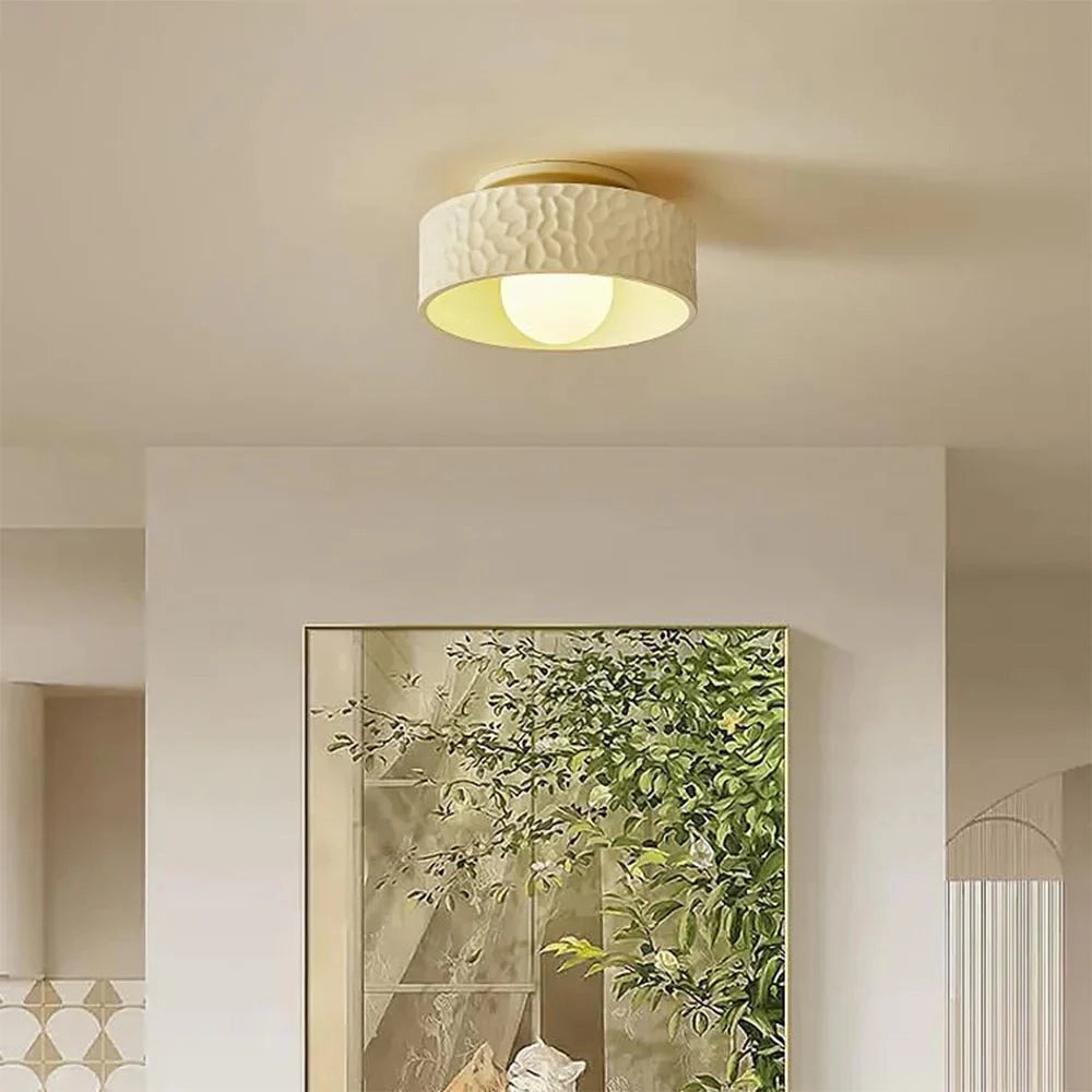 Modern textured circular LED ceiling light fixture mounted on a white ceiling with a flush design, featuring a sleek, minimalist look in a contemporary interior setting. The surrounding space includes elements of interior design such as a plant and wooden accents against a light-colored wall, creating a harmonious and stylish atmosphere.