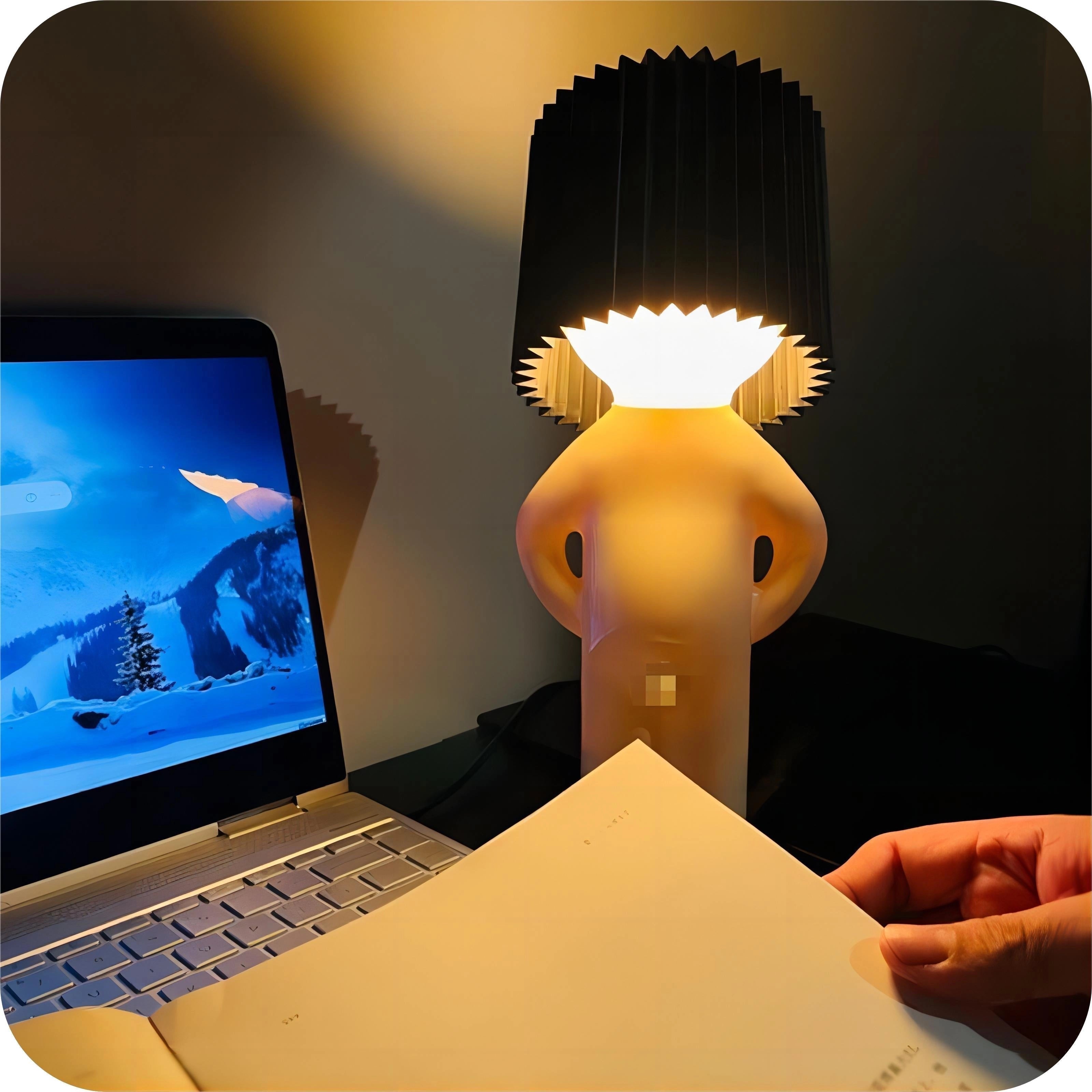 A black novelty human-shaped table lamp placed on a desk, emitting a warm glow and serving as a quirky designer accent lighting piece. The lamp features a unique human form, adding a playful and artistic touch to the workspace.