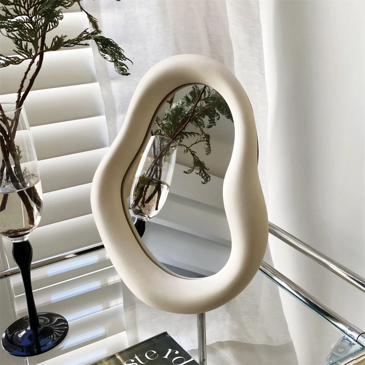 Organic asymmetrical mirror with a modern minimalist design, showcasing a reflection of a room with a window, potted plant, and art pieces, contributing to a stylish interior decor setup.