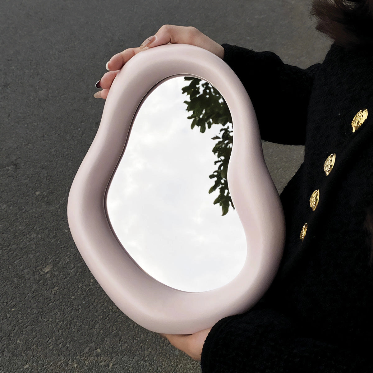 Organic asymmetrical mirror with a modern minimalist design featuring a pink frame, displayed in a vanity setting with a blurred reflection of a person making a gesture.