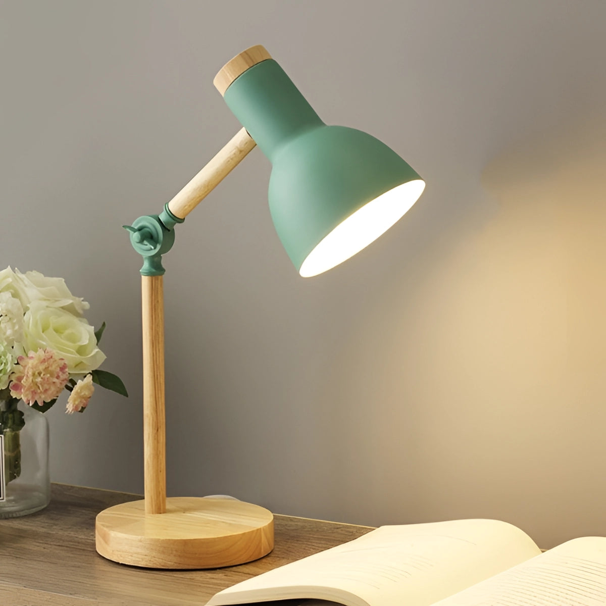 Scandinavian wooden adjustable LED desk lamp in retro style featuring a green finish and a switch for three color settings, placed on a table next to a small plant and a rose.