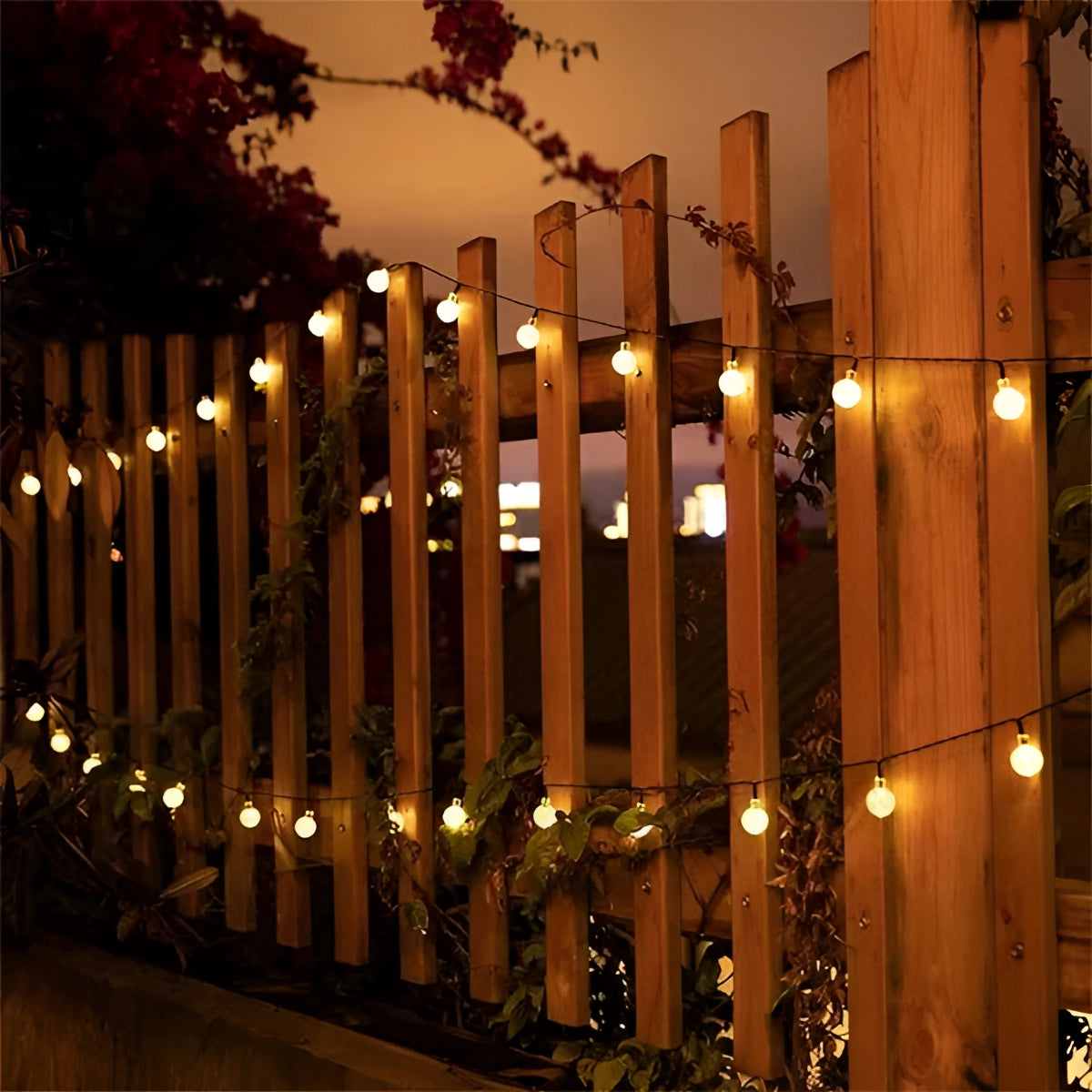 String lights with solar crackled glass globes hanging on a wooden fence in an outdoor setting, with trees and a clear sky visible in the background.