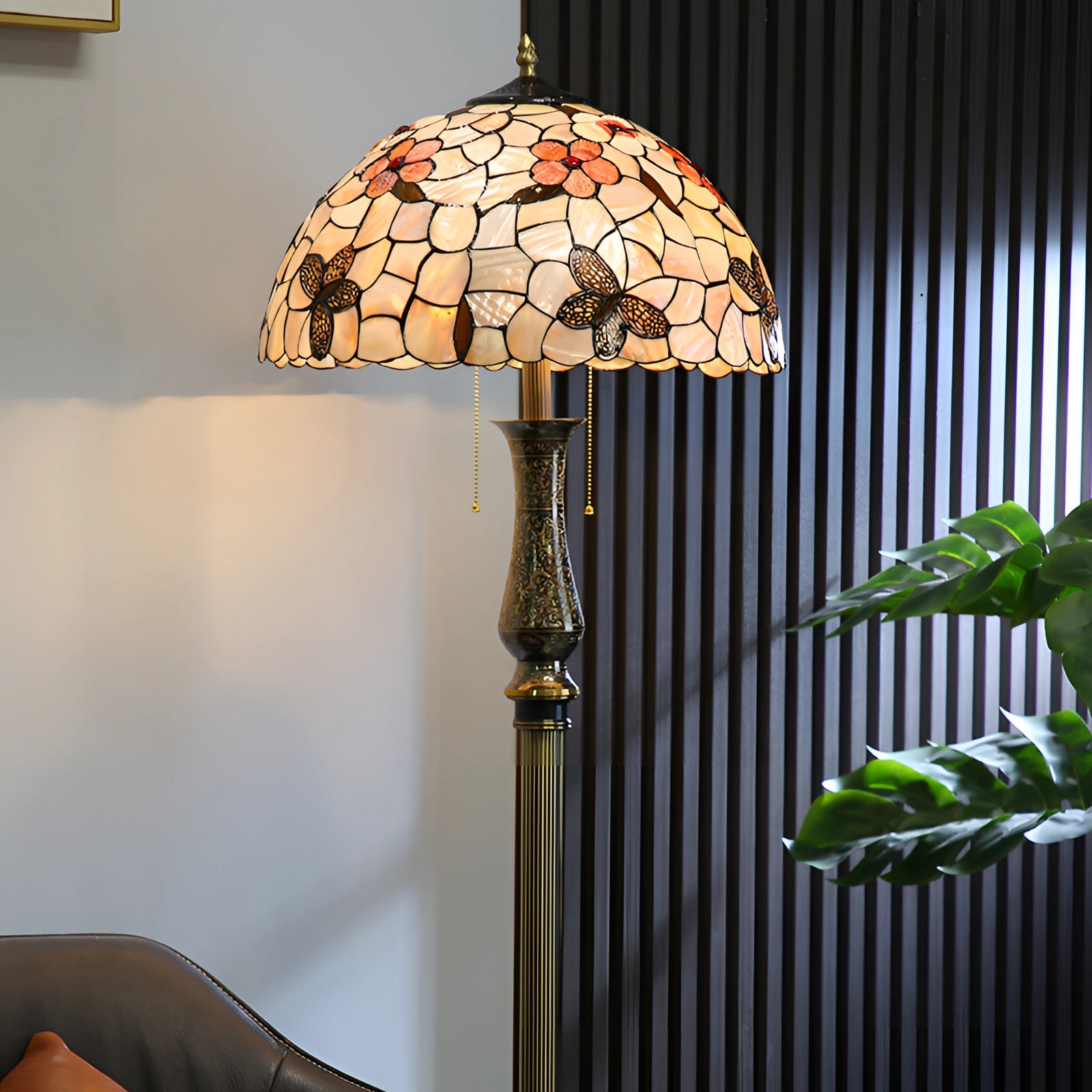 Tiffany-style stained glass floor lamp with dragonfly design shade, featuring a brass pole, positioned indoors near a window with plants in the background.