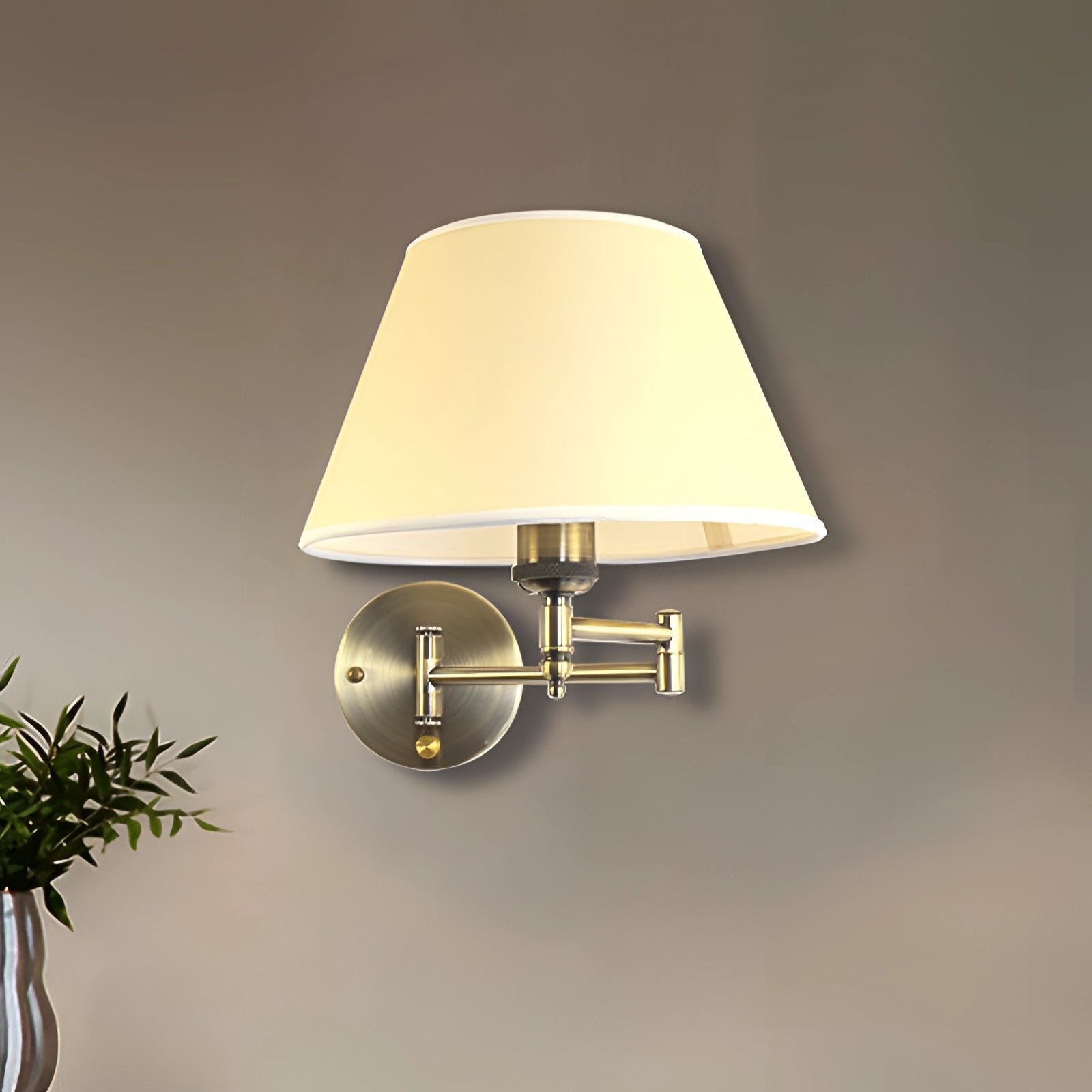 Vintage brass wall sconce with pleated beige shade and adjustable arm mounted on a wall above a wooden table, surrounded by houseplants and interior decor elements.