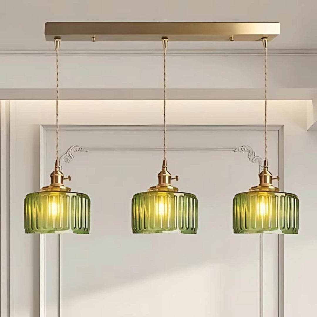 Vintage green glass pendant light with three long ribbed glass heads and antique brass fittings, hanging in a bright room with wooden shelving and window in the background.