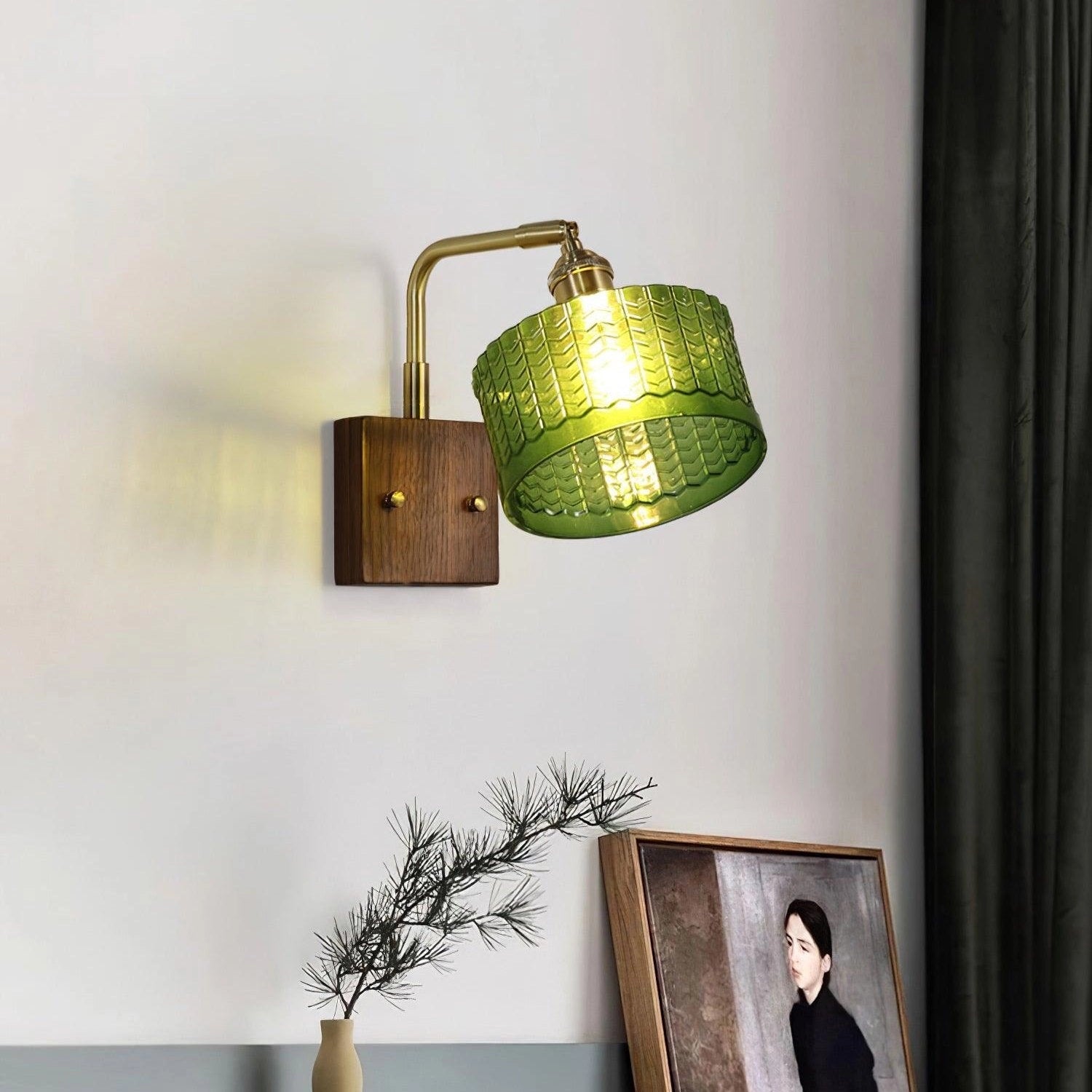 Vintage green glass sconce with a brass arm featuring a fish scale pattern, positioned against a wooden backdrop with decorative elements including a plant and picture frame nearby.